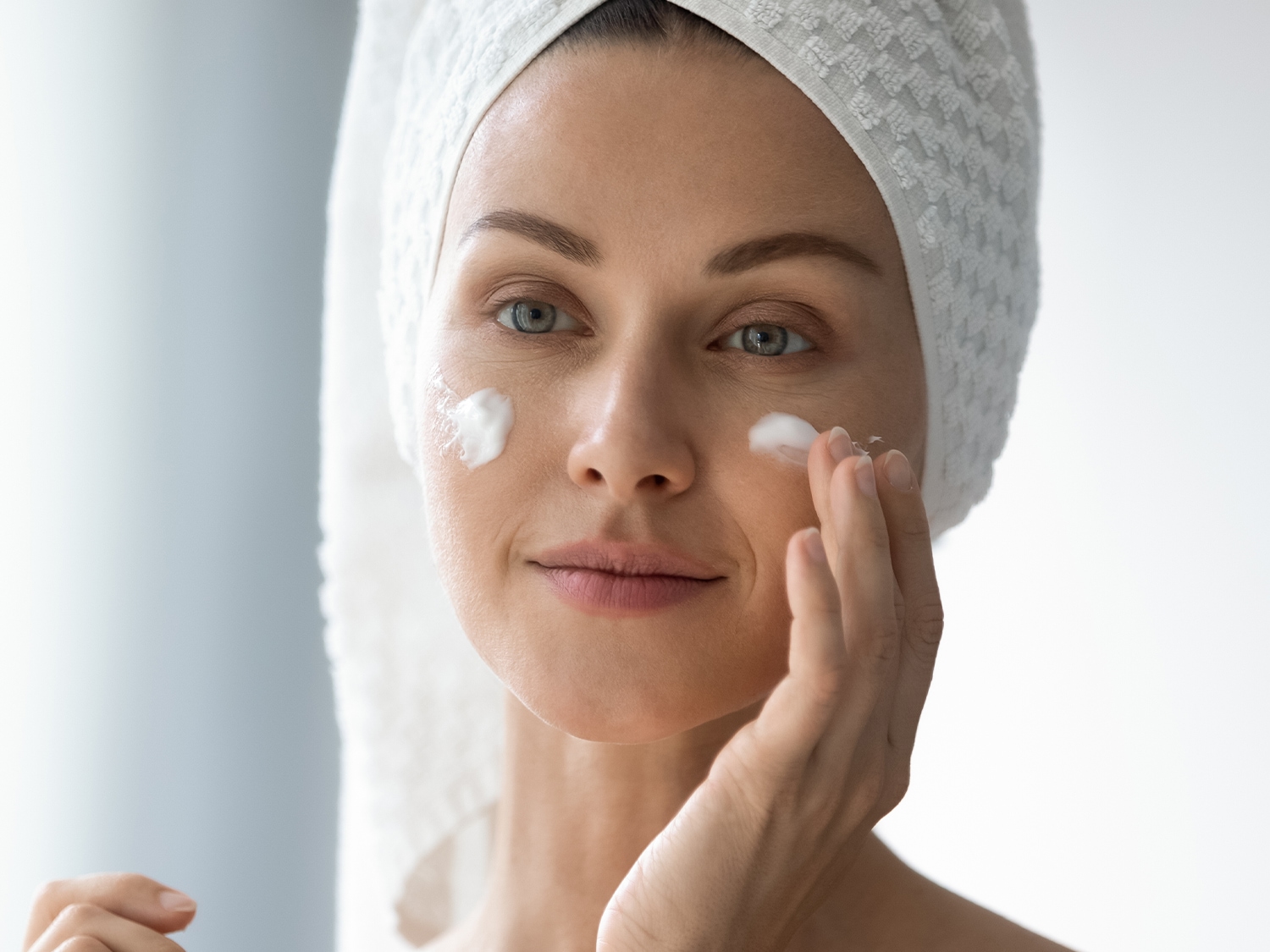 Primo piano di una donna con un asciugamano bianco in testa che applica una crema bianca sul viso, per una pelle idratata e luminosa.