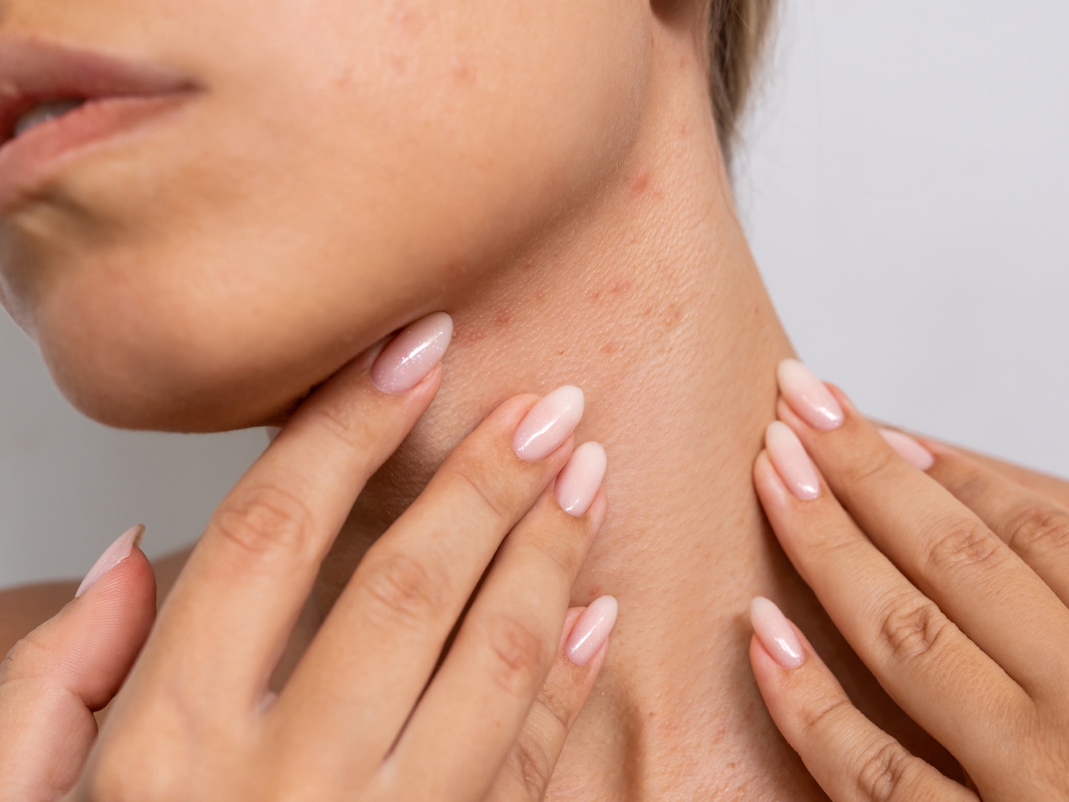 Primo piano di una donna che tocca il collo con le mani, mostrando la texture della pelle e le unghie rosa lucide.