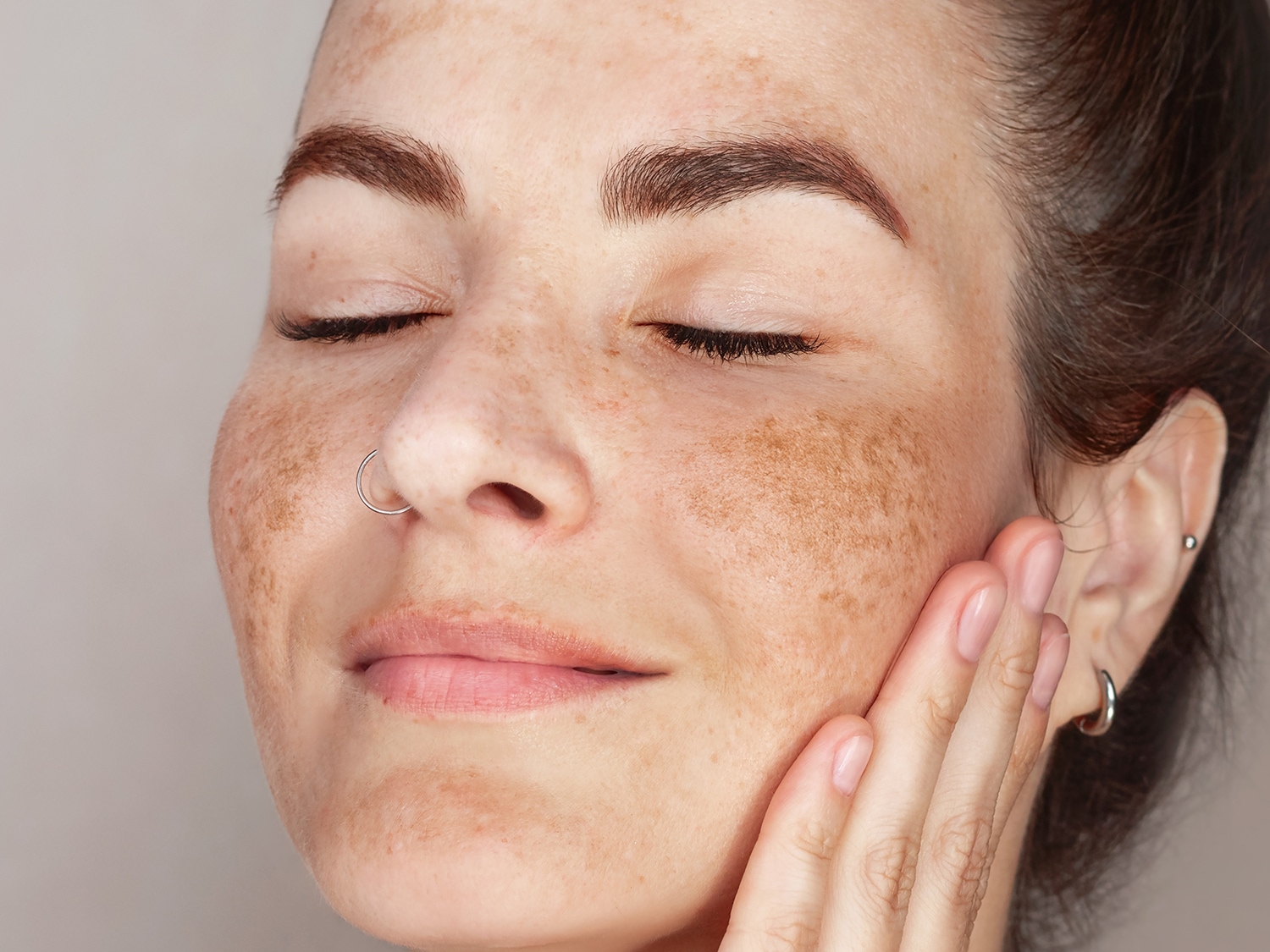 Primo piano di una donna con lentiggini, occhi chiusi e un piercing al naso, che tocca delicatamente la sua guancia con la mano.