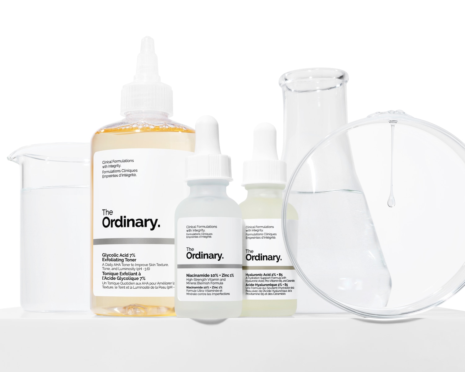 Drei The Ordinary Produkte stehen neben Laborelementen auf einer weißen Platte vor weißem Hintergrund.