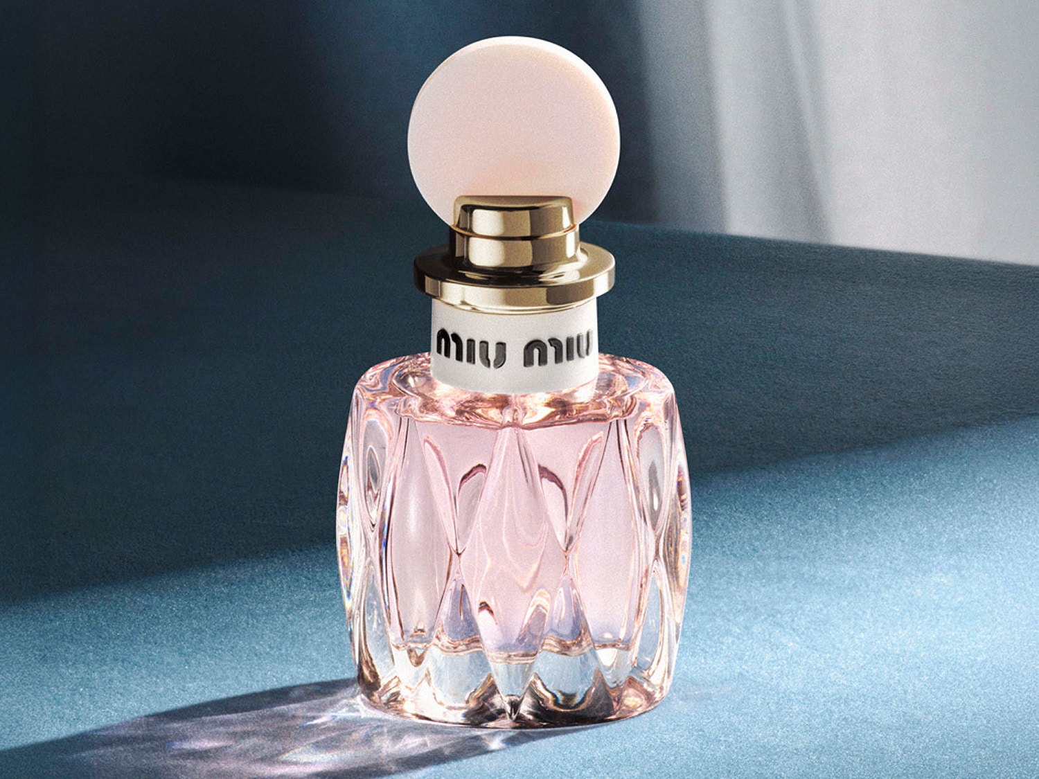 Butelka perfum Miu Miu z różowym płynem w fasetowanej, przezroczystej butelce, zwieńczona okrągłą, jasnoróżową nakrętką i złotym pierścieniem.