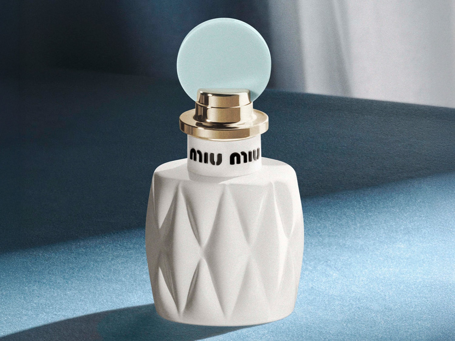 Biały flakon perfum Miu Miu z fasetowaną powierzchnią i jasnoniebieskim okrągłym korkiem, otoczonym złotym pierścieniem, stoi na niebieskim tle.