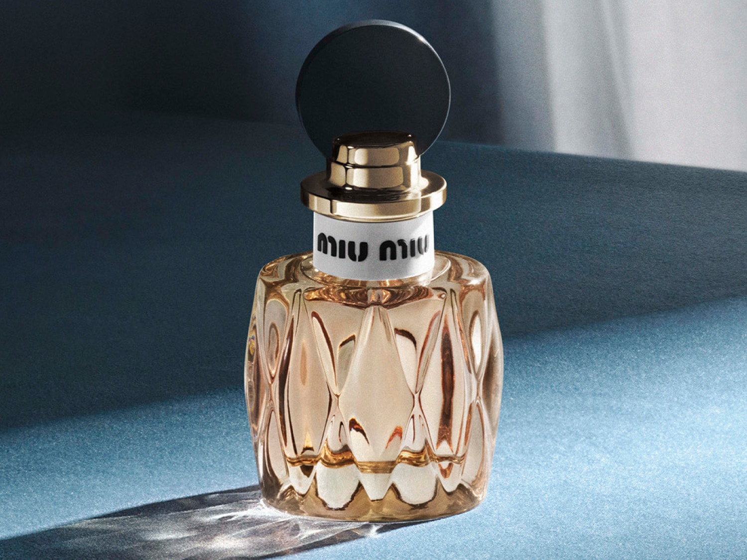 Bursztynowy flakon perfum Miu Miu z geometrycznymi fasetami i czarnym korkiem na niebieskim tle emanuje luksusem i wyrafinowaniem.