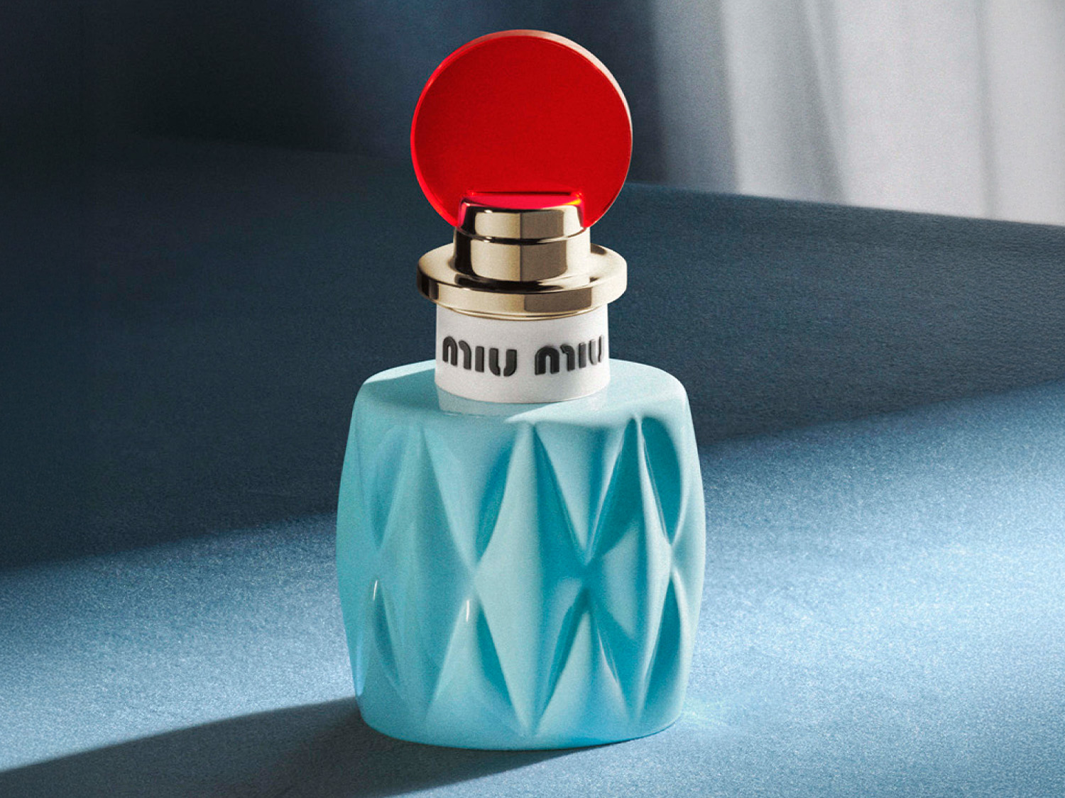 Błękitny flakon perfum Miu Miu z fasetowaną powierzchnią i czerwonym korkiem na tle niebieskiego materiału.