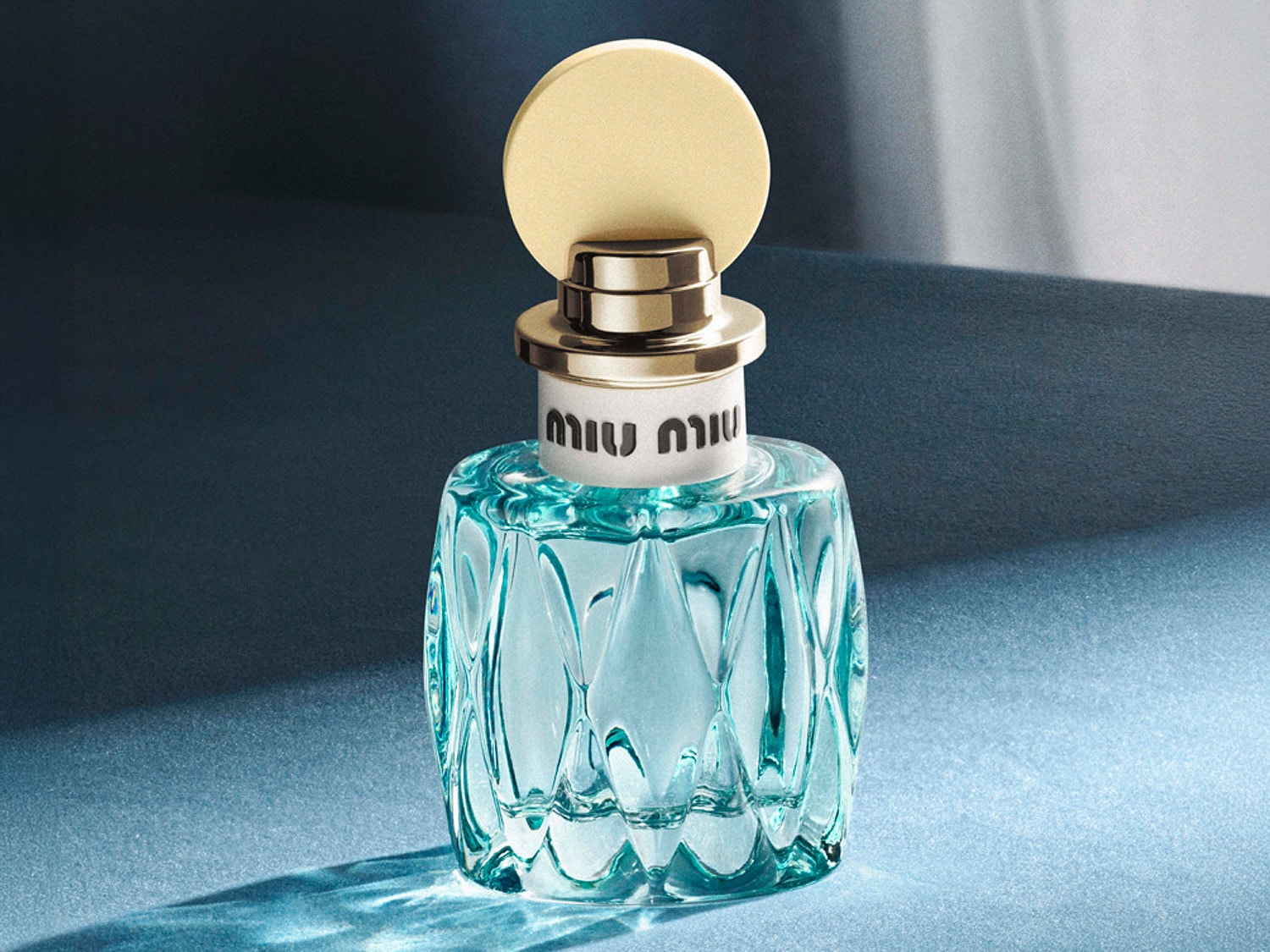 Błękitny flakon perfum Miu Miu z fasetowanego szkła, z kremowozłotą nakrętką w kształcie dysku, emanuje świeżością i elegancją.