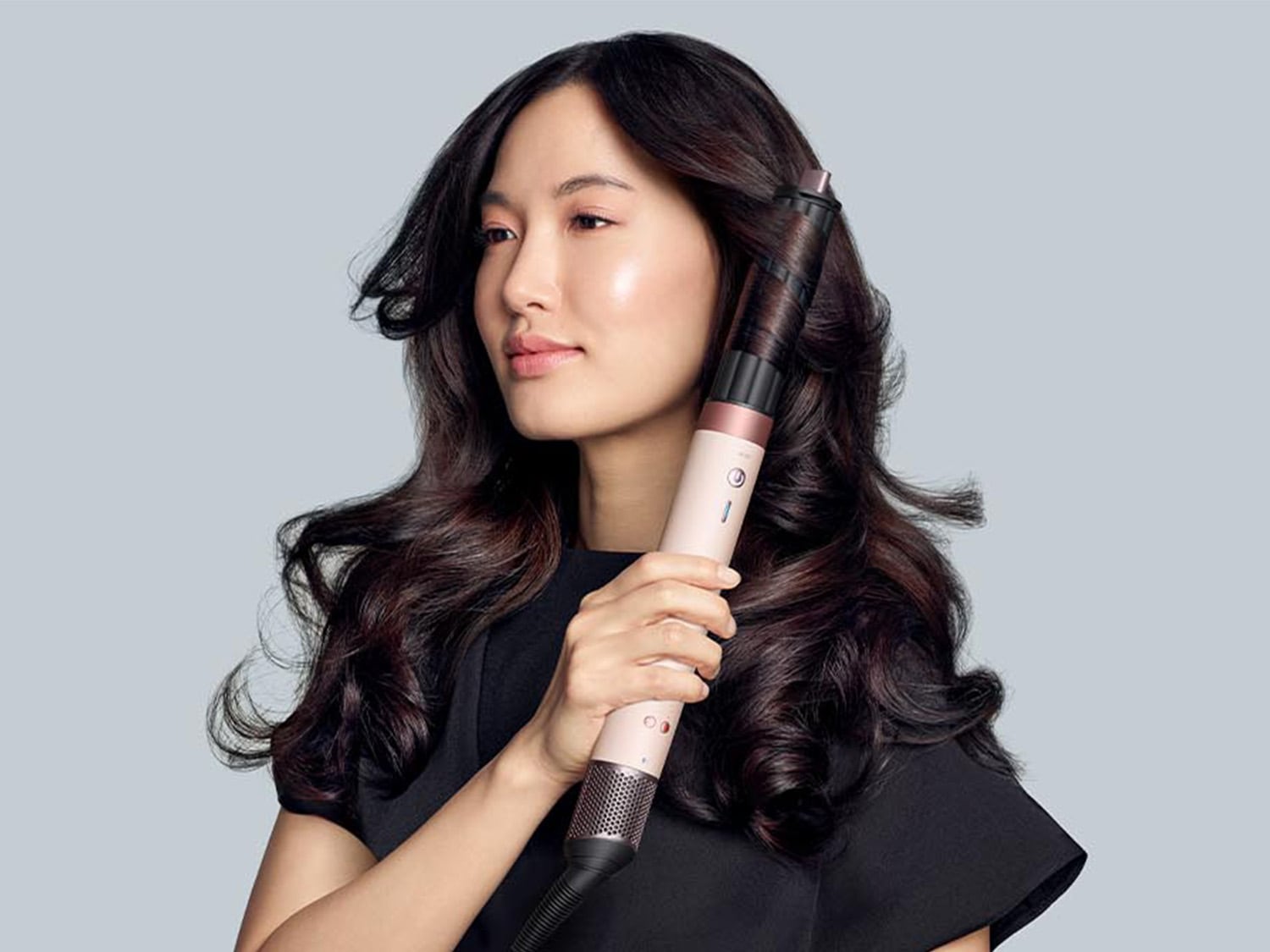 Eine Frau mit dunklen, gewellten Haaren hält einen Lockenstab in der Hand, um ihre Haare zu stylen.