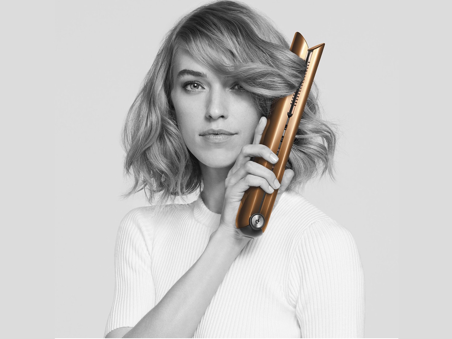 Eine Frau mit gewelltem, schulterlangem Haar hält ein goldenes Glätteisen an ihr Gesicht.