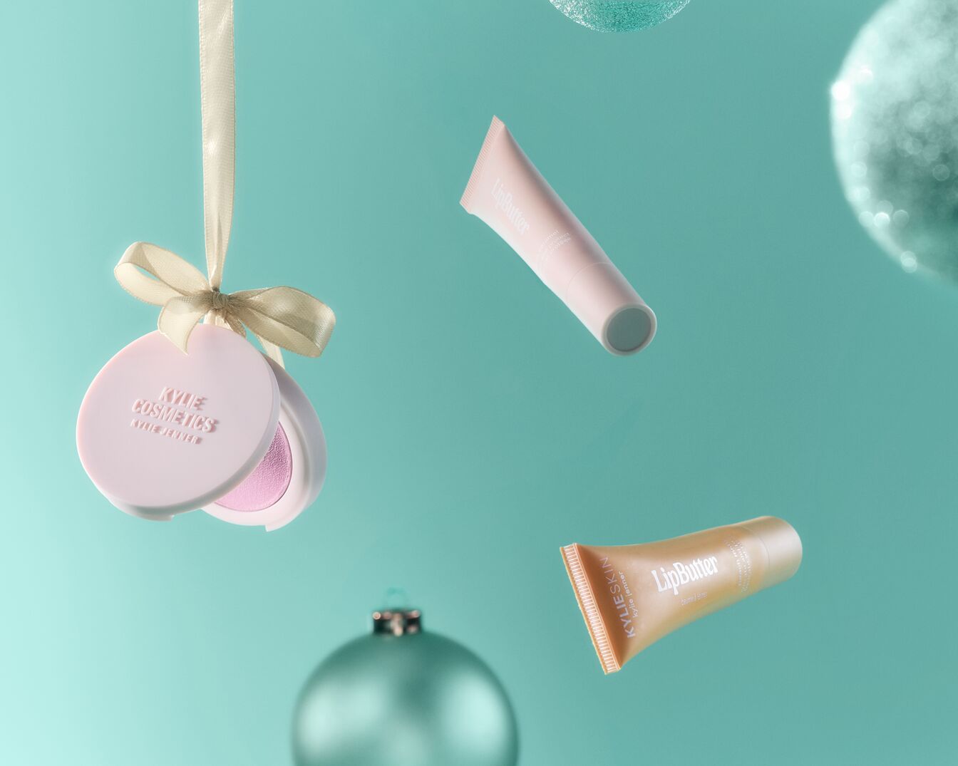 Na turkusowym tle unoszą się błyszczące ozdoby, puder Kylie Cosmetics zawieszony na wstążce oraz tubki LipButter w odcieniach różu i złota.