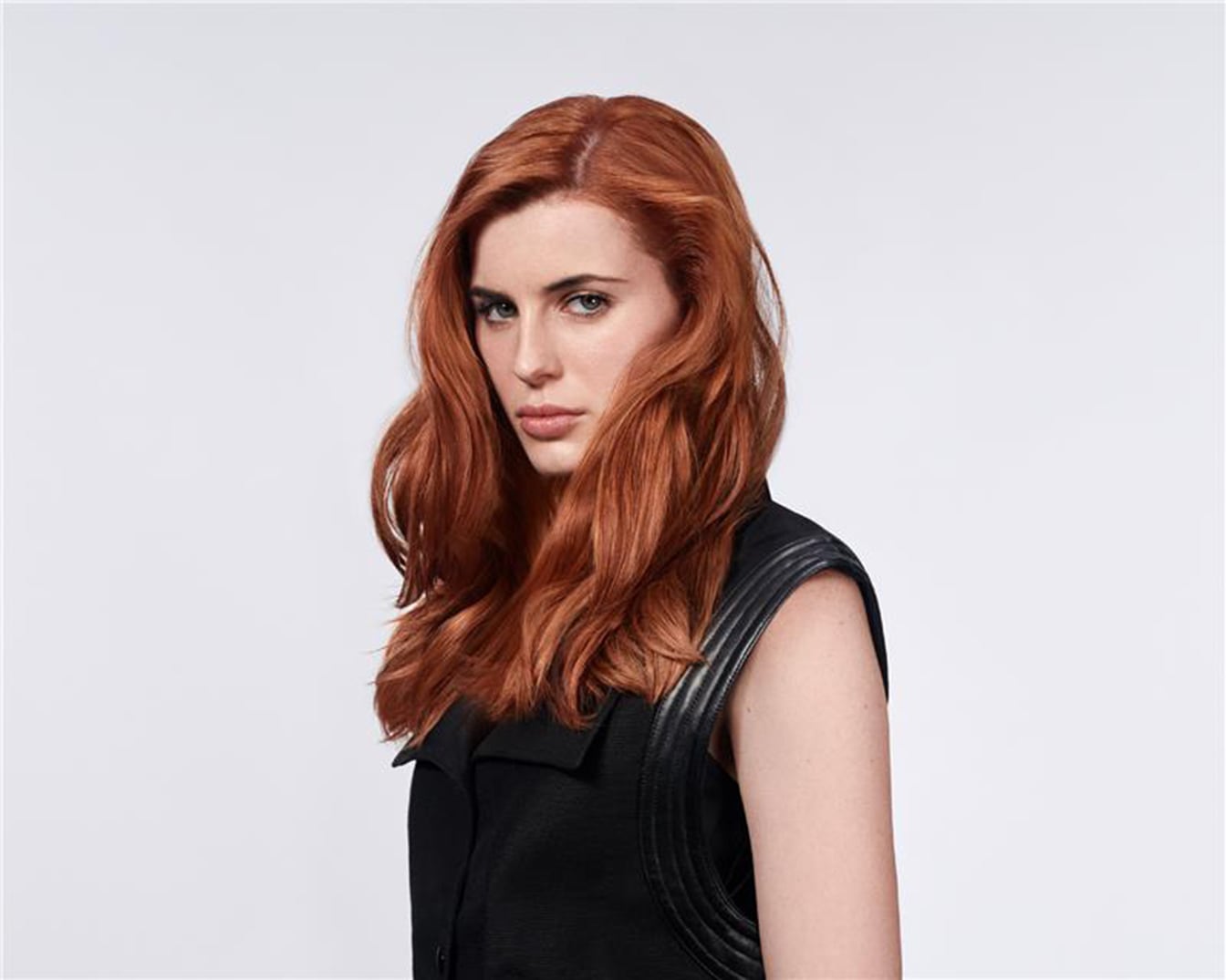 Portrait d'une femme aux cheveux roux ondulés, portant un haut noir sans manches, sur un fond blanc uni.