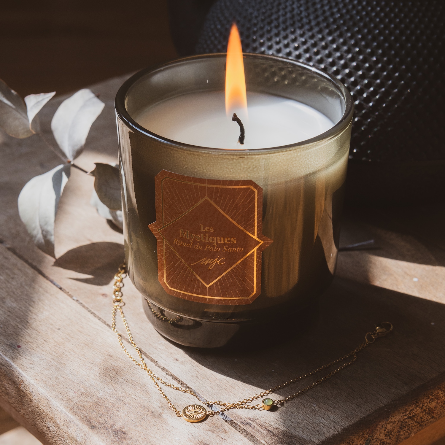 Bougie parfumée allumée dans un récipient en verre brun clair avec une étiquette marron et un bracelet doré en dessous.