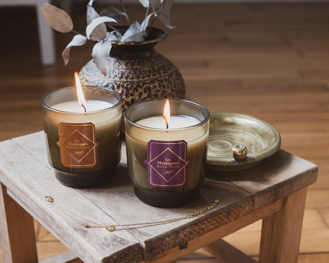 Deux bougies parfumées "Les Mystiques" sont posées sur un tabouret en bois, avec un vase décoratif et un collier doré à proximité, créant une ambiance chaleureuse et invitante.