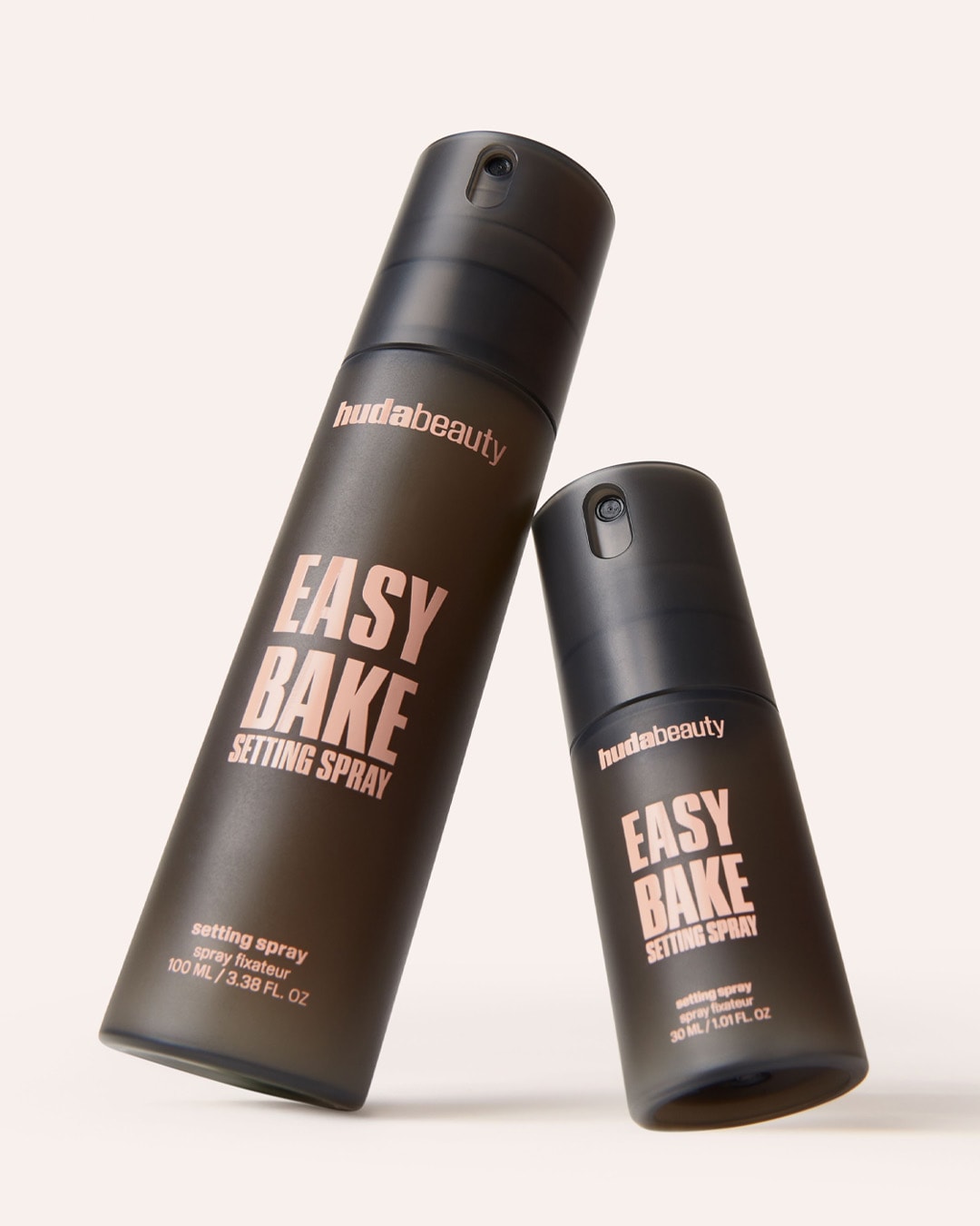 Zwei Sprühflaschen Huda Beauty Easy Bake Setting Spray, eine 100 ml und eine 30 ml, beide in einem matten, dunklen Farbton mit roségoldener Schrift.