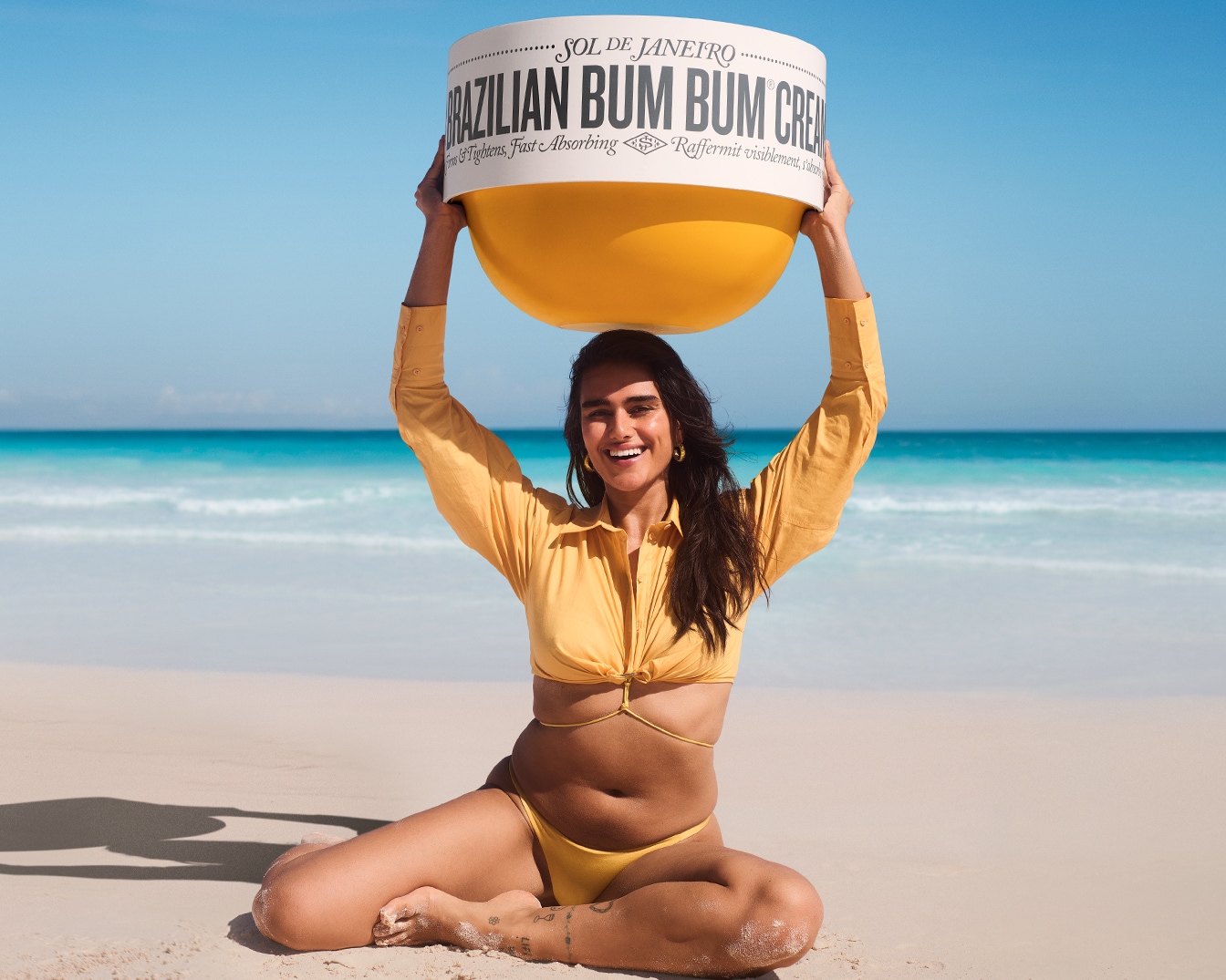 Eine Frau sitzt am Strand und hält eine übergroße Brazilian Bum Bum Cream von Sol de Janeiro über ihrem Kopf, die deine Haut strafft und schnell einzieht.