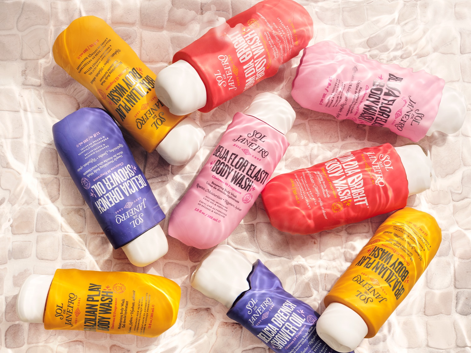 Eine Sammlung von Duschgelen und Duschölen von Sol de Janeiro in verschiedenen Farben und Düften, darunter Brazilian Play, Delicia Drench und Bom Dia Bright, die auf einer hellen, gefliesten Oberfläche im Wasser liegen.
