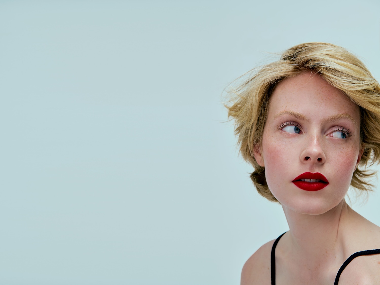 Nahaufnahme einer Frau mit blonden, gewellten Haaren, roten Lippen und Sommersprossen, die mit einem trägerlosen schwarzen Oberteil bekleidet ist und nach links blickt.