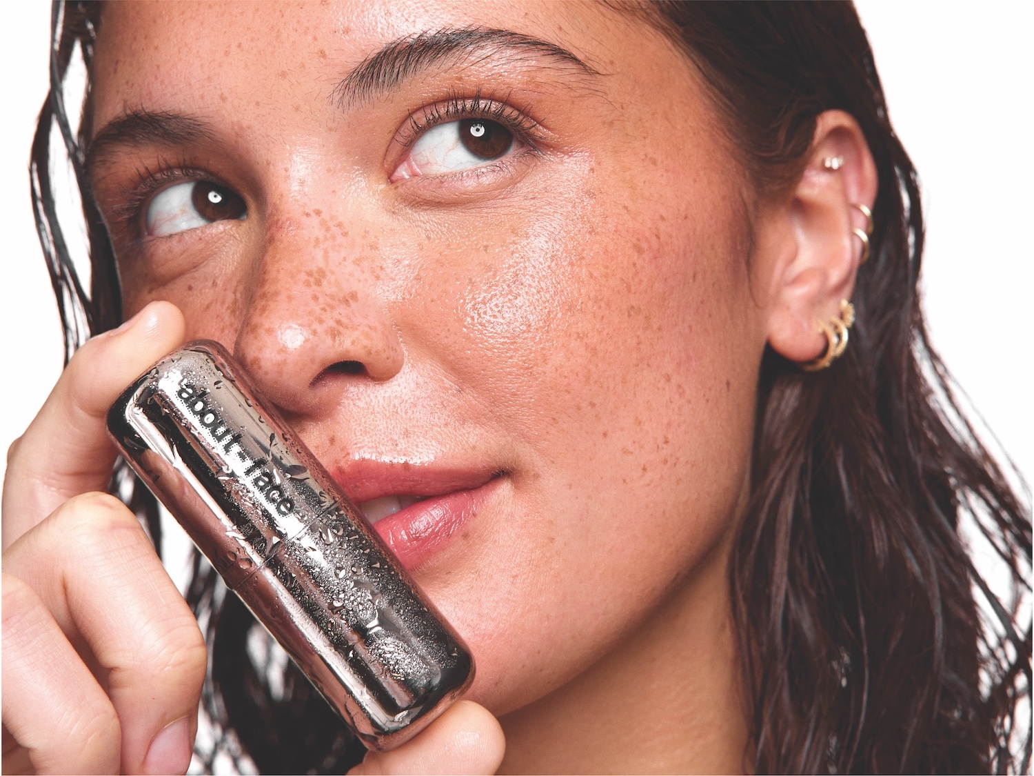 Une femme souriante tient un tube argenté de About Face, sa peau éclatante et ses lèvres brillantes captent la lumière.