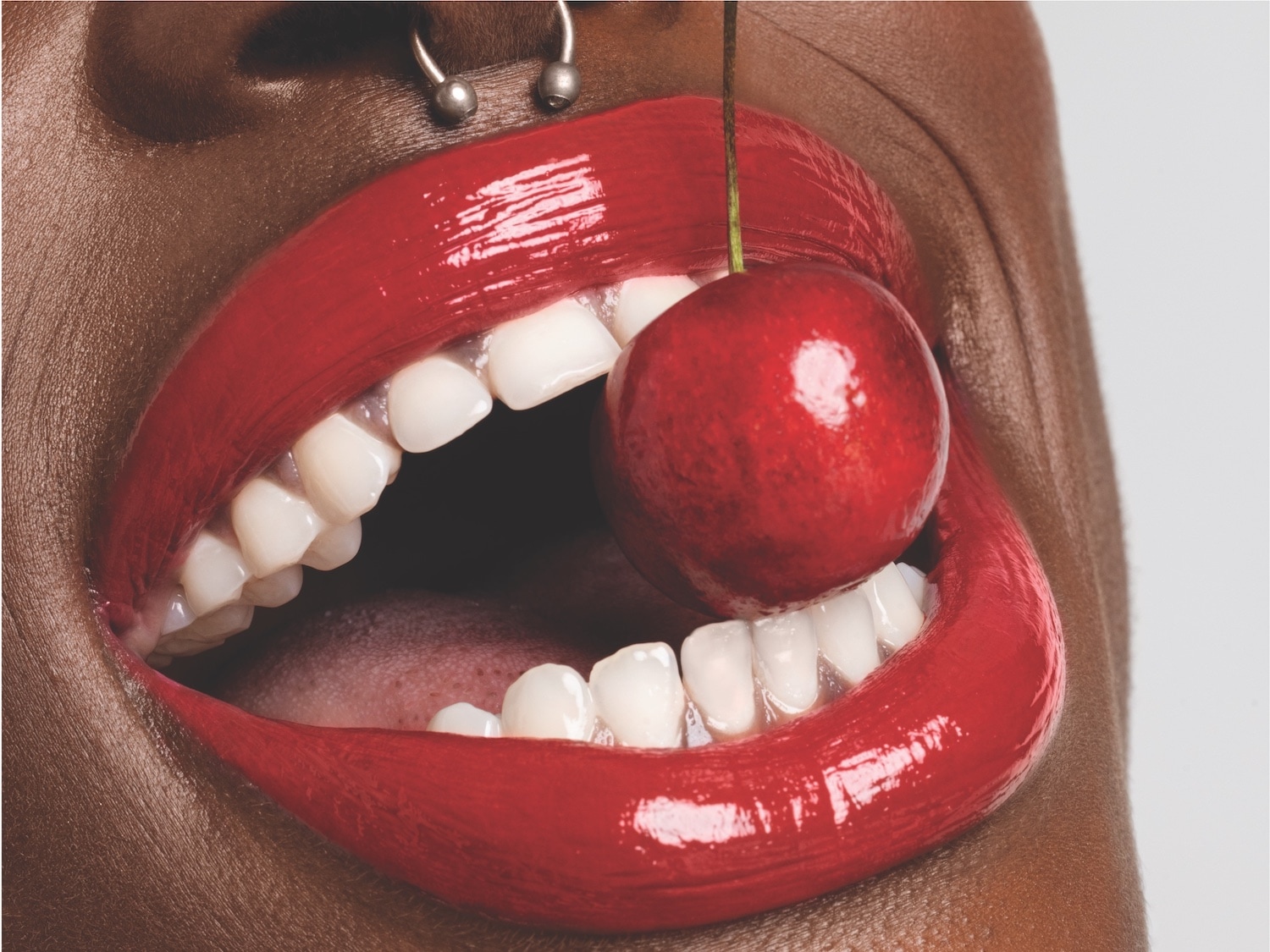 Nahaufnahme von einem Mund mit glänzend rotem Lippenstift und einer Kirsche zwischen den Zähnen.