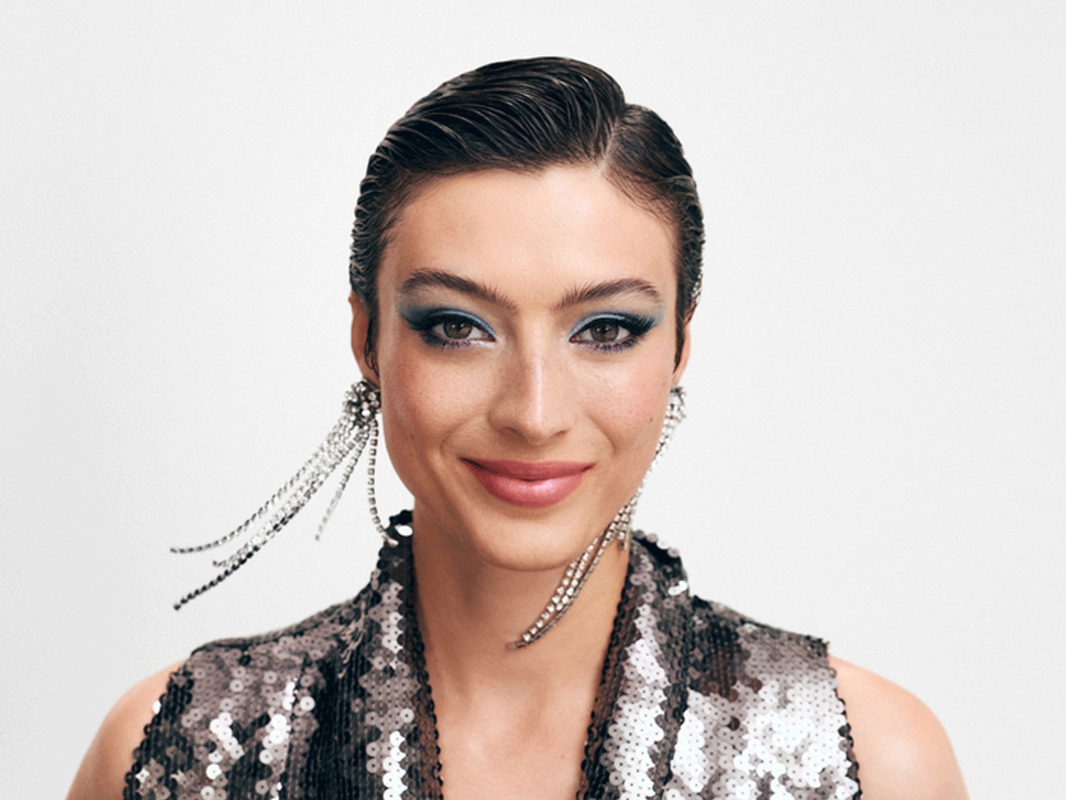 Nahaufnahme einer Frau mit glatt zurückgekämmtem, dunklem Haar, blauem Lidschatten und einem glitzernden Paillettenoberteil, dazu baumelnde Ohrringe.