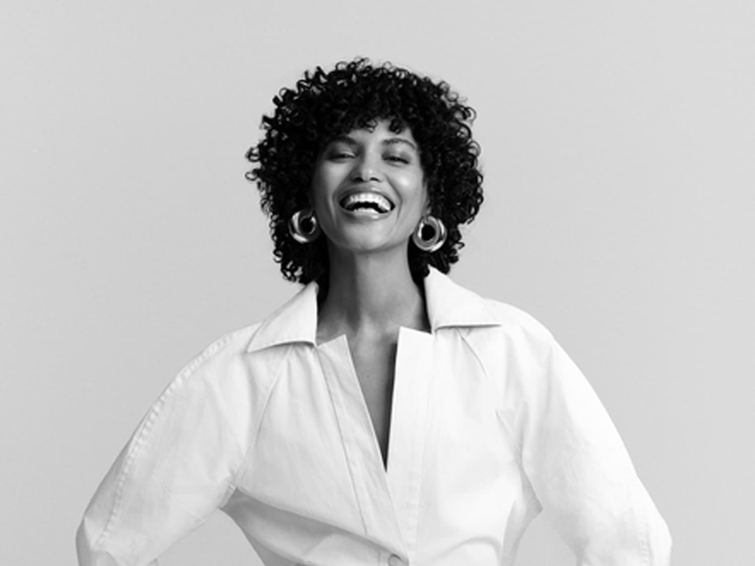 Schwarz-Weiß-Foto einer lachenden Frau mit lockigem Haar, die ein weißes Hemd und große Creolen trägt.