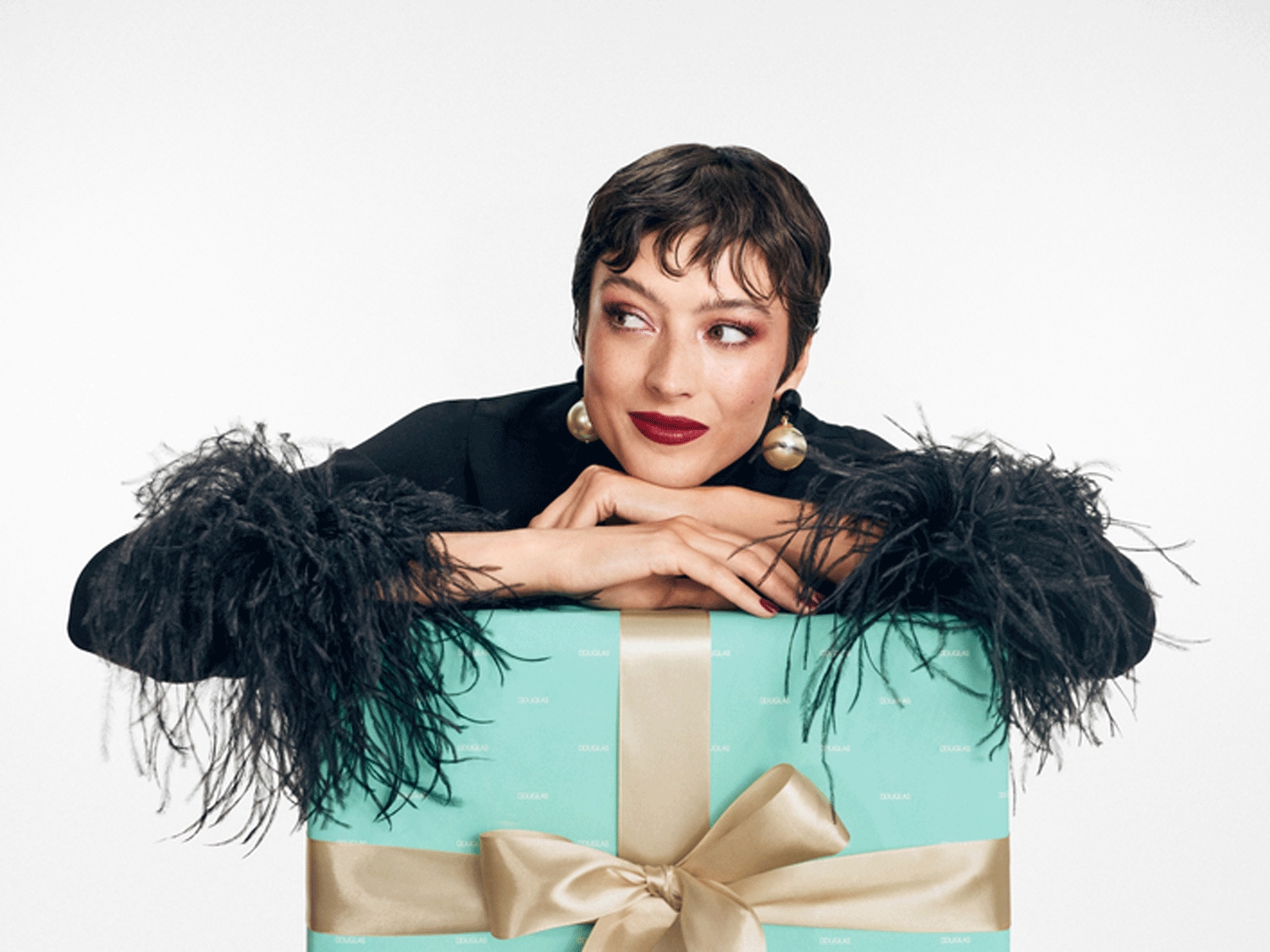 Eine Frau mit dunklem Haar und Ohrringen posiert mit einem hellblauen Geschenk, das mit einer goldenen Schleife verziert ist.