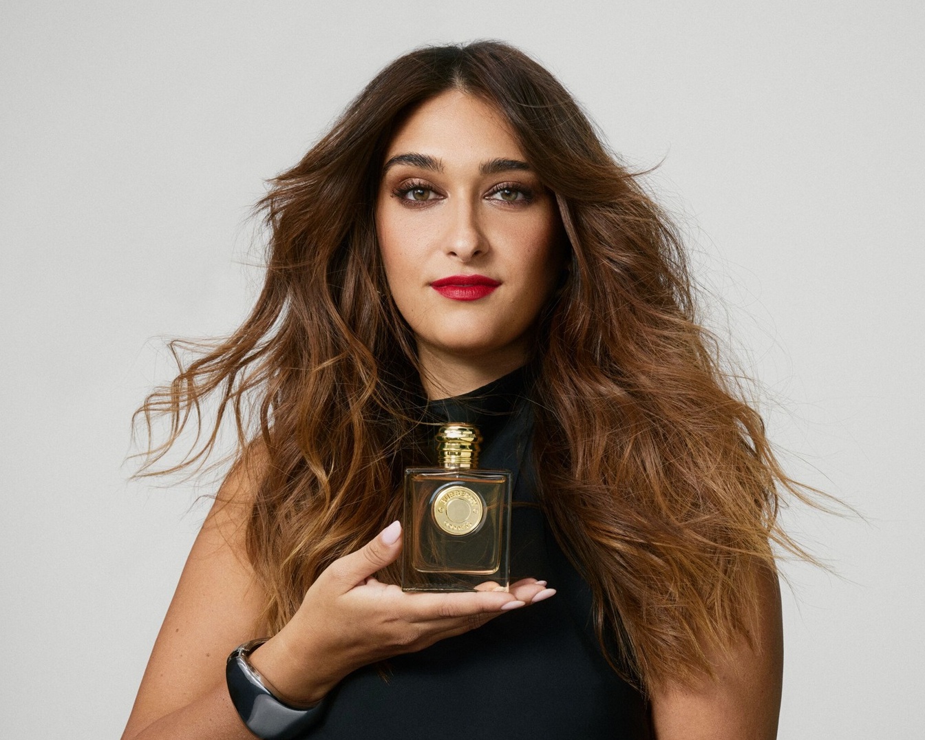Une femme aux longs cheveux bruns ondulés tient un flacon de parfum carré transparent avec un bouchon doré, portant un haut noir et un rouge à lèvres rouge vif.