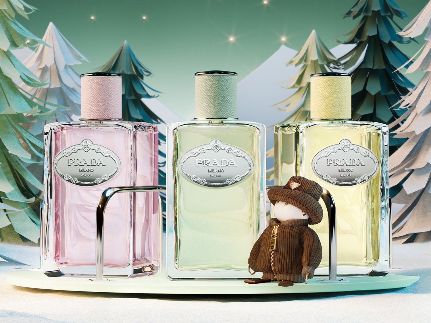 Na tle zimowej scenerii, trzy flakony perfum Prada o pastelowych odcieniach różu, zieleni i żółci, prezentowane na eleganckim stojaku, z uroczą figurką w zimowym ubranku.