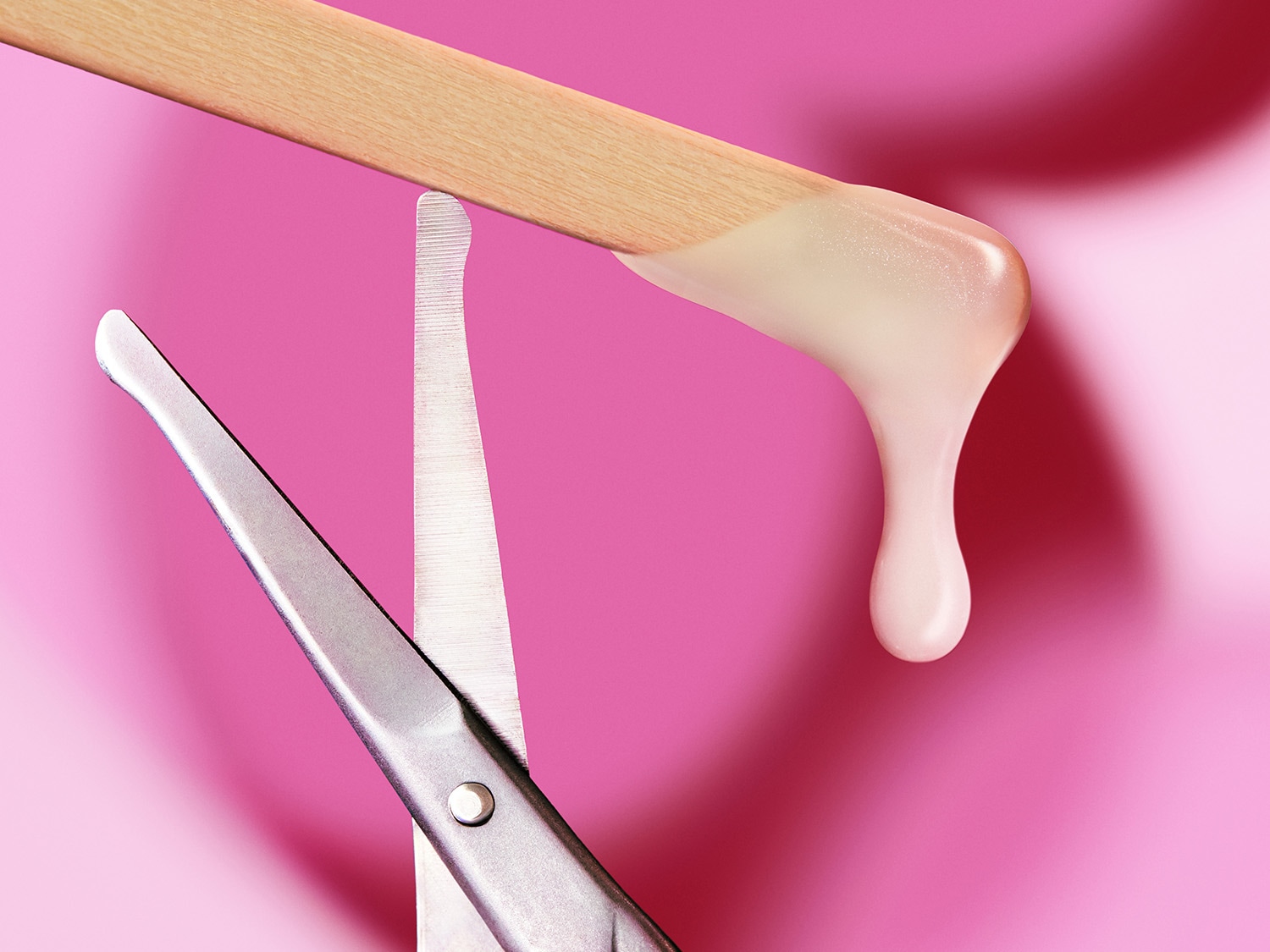 Nahaufnahme von Wachs auf einem Holzspatel, der über einer Schere auf einem rosa Hintergrund gehalten wird.