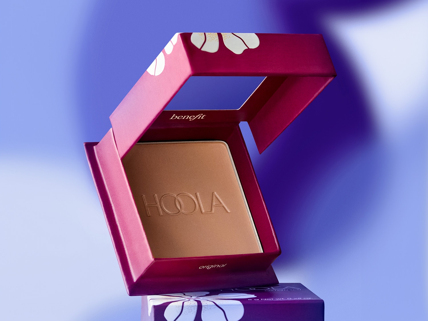 Benefit Hoola Bronzer in einer rosafarbenen, geöffneten Schachtel mit weißen Blumen auf blauem Hintergrund.