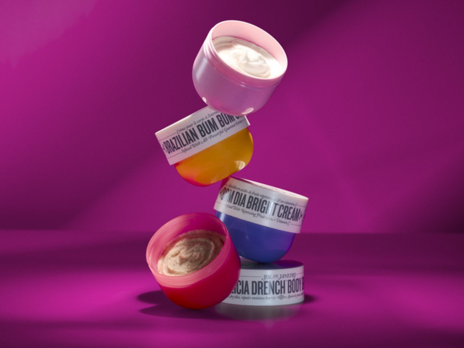 Stapel van Sol de Janeiro crèmes, waaronder Brazilian Bum Bum, Bom Dia Bright Cream en de Coco Cabana Drench Body Lotion, elk met een unieke kleur dop, tegen een levendige magenta achtergrond.