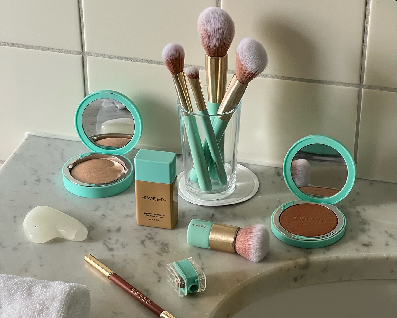 Na marmurowej umywalce leżą kosmetyki Sweed w turkusowych opakowaniach, w tym pędzle do makijażu w szklance, pudry, podkład, konturówka i temperówka.