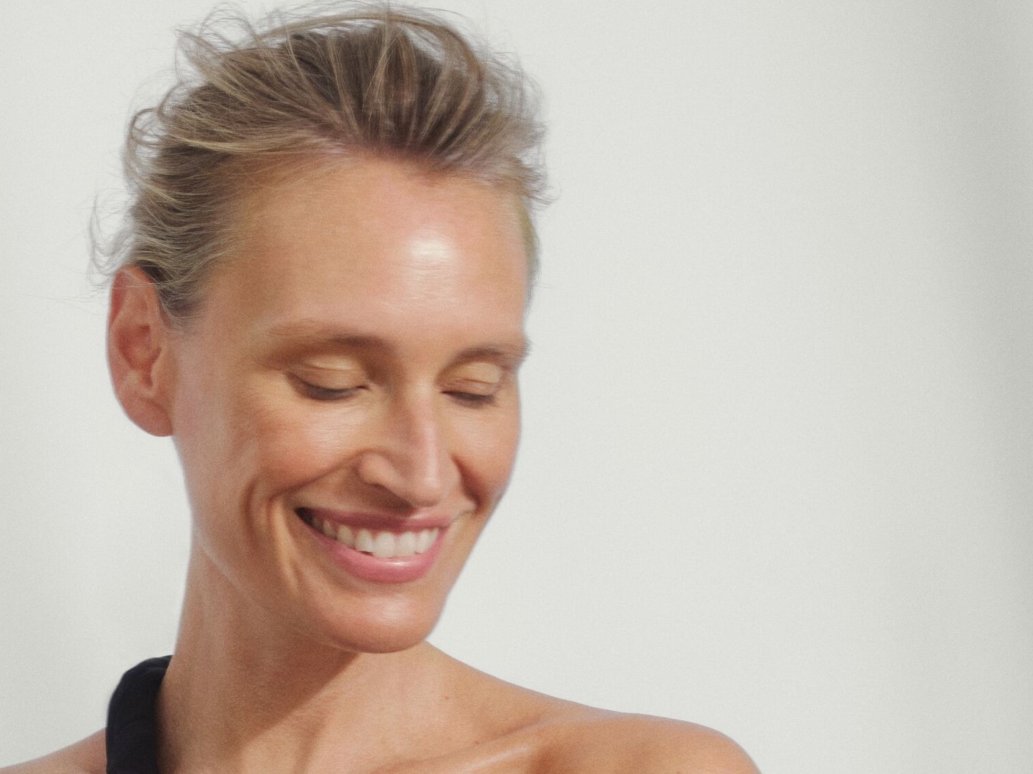 Nahaufnahme einer Frau mit blonden Haaren, die lächelt und ihre Augen geschlossen hat, während sie in einem hellen, neutralen Raum steht.