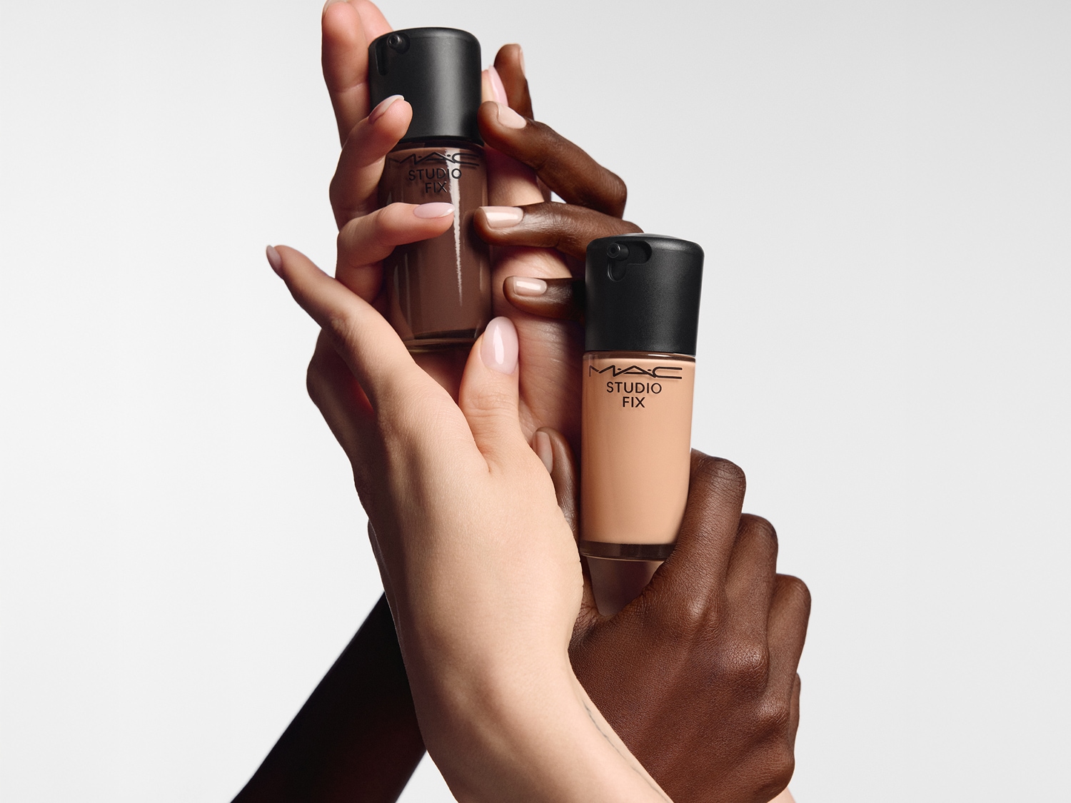 Mehrere Hände präsentieren die MAC Studio Fix Foundation in verschiedene Hauttönen in Glasflaschen mit schwarzem Deckel.