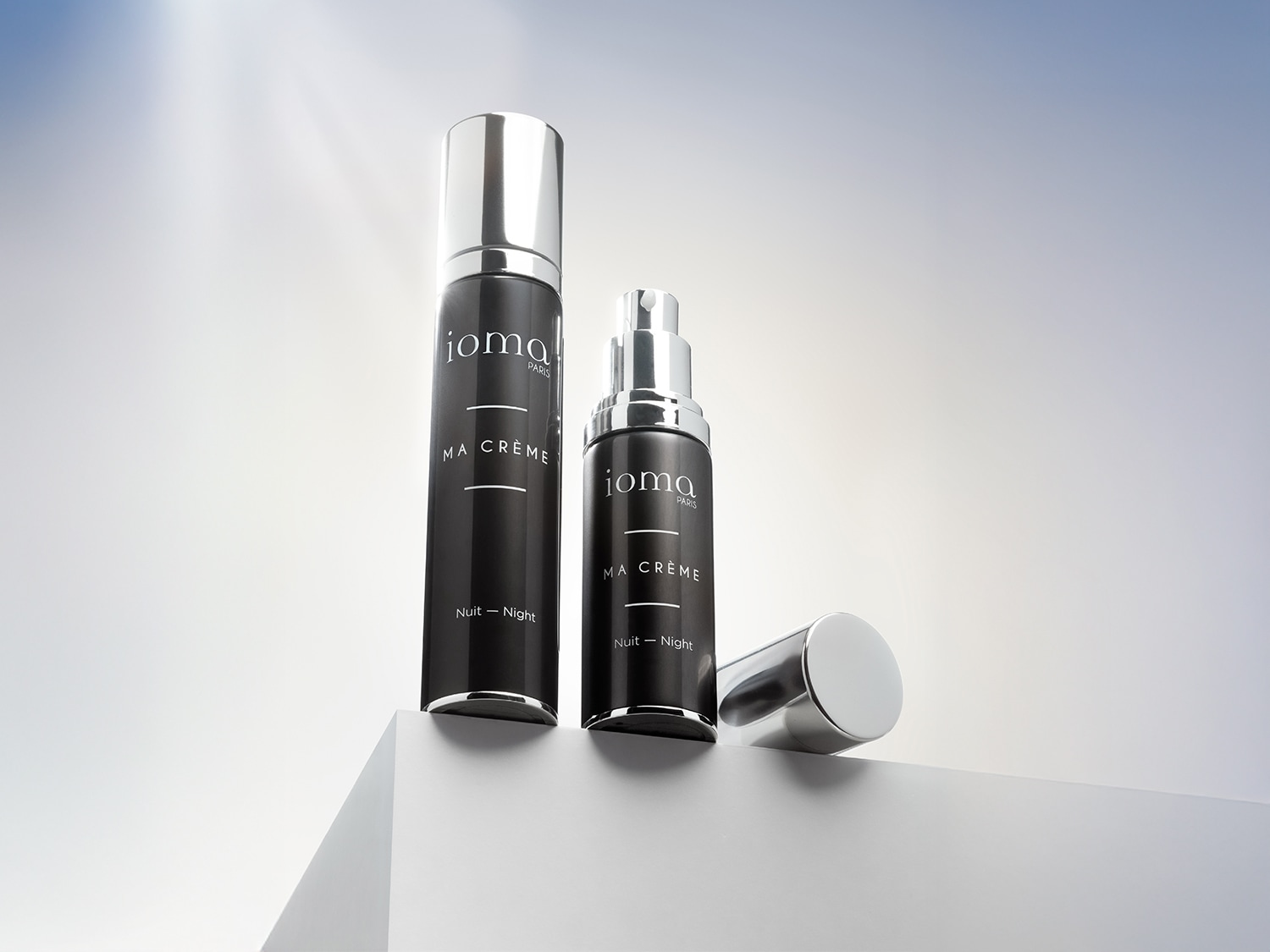 Ecco due flaconi di crema da notte Joma Paris Ma Crème, uno alto e sottile e l'altro più corto e tozzo, entrambi con tappi argentati lucidi.