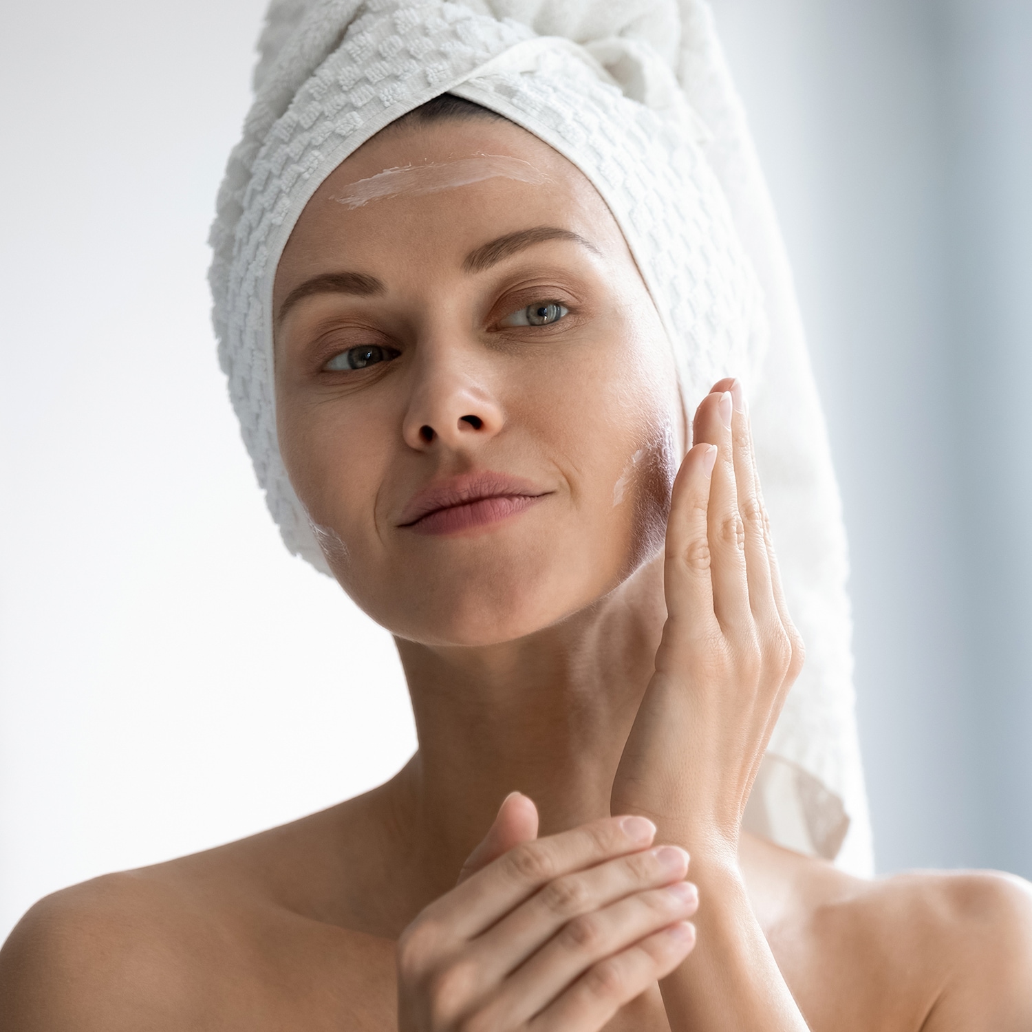 Primo piano di una donna con un asciugamano bianco in testa che si applica una crema bianca sul viso con la mano.