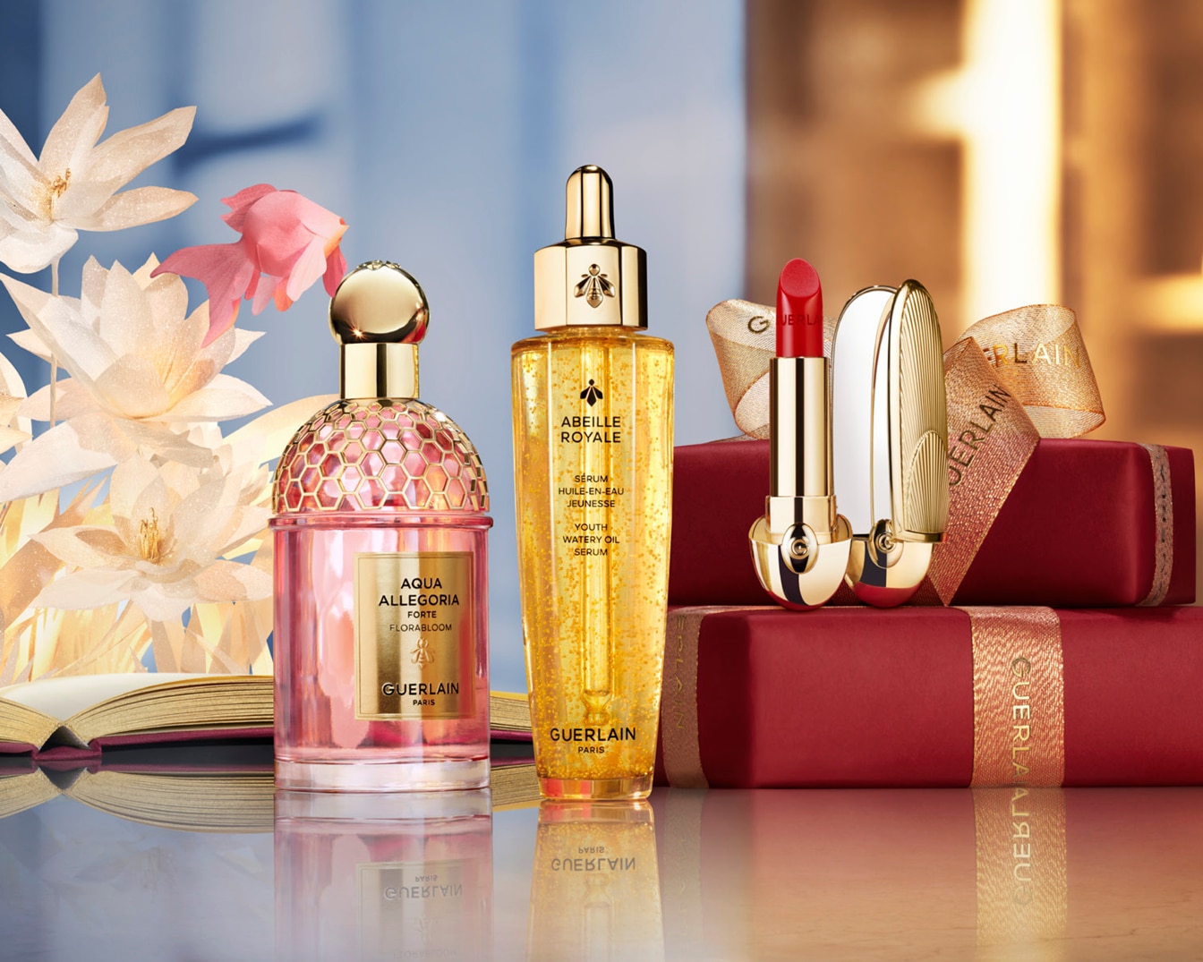 A Guerlain Aqua Allegoria Forte Florabloom parfüm rózsaszín üvegben, az Abeille Royale Youth Watery Oil szérum arany színben, és egy piros rúzs arany csomagolásban, piros ajándékdobozokon.