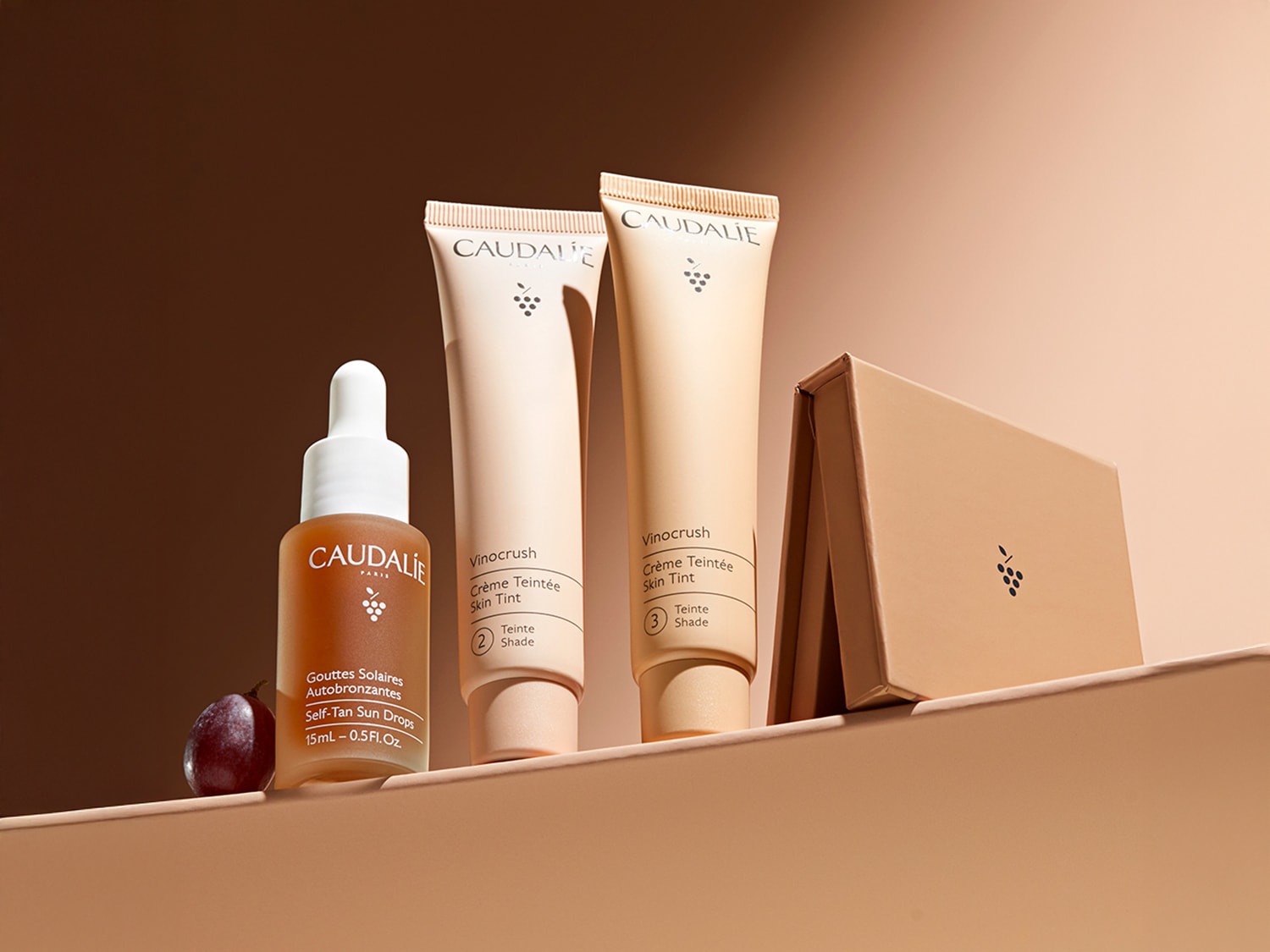 Eine Kollektion von Vinocrush-Hauttönungscremes von Caudalie in den Farbtönen 2 und 3, Selbstbräunungstropfen in einer bernsteinfarbenen Glasflasche mit Pipette und eine quadratische Puderdose.