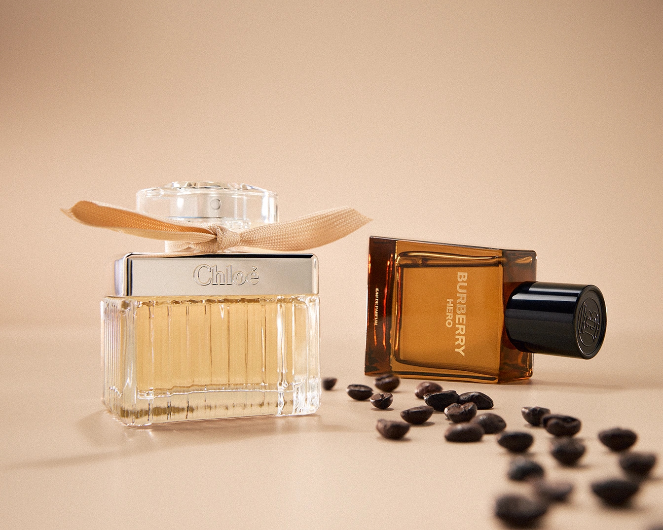 Zwei Parfümflaschen, eine von Chloé mit einer Schleife und eine von Burberry Hero, liegen auf einem beigen Untergrund, umgeben von Kaffeebohnen.