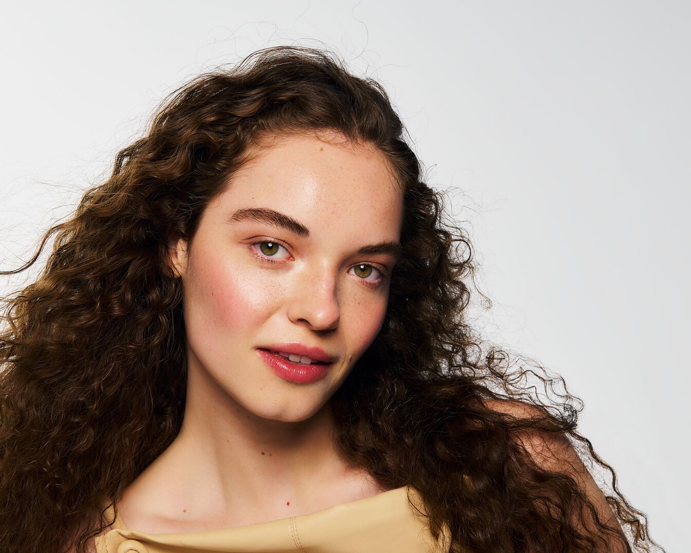 Portrét mladé ženy s vlnitými hnědými vlasy, jemným make-upem a zelenýma očima, která se dívá do objektivu.