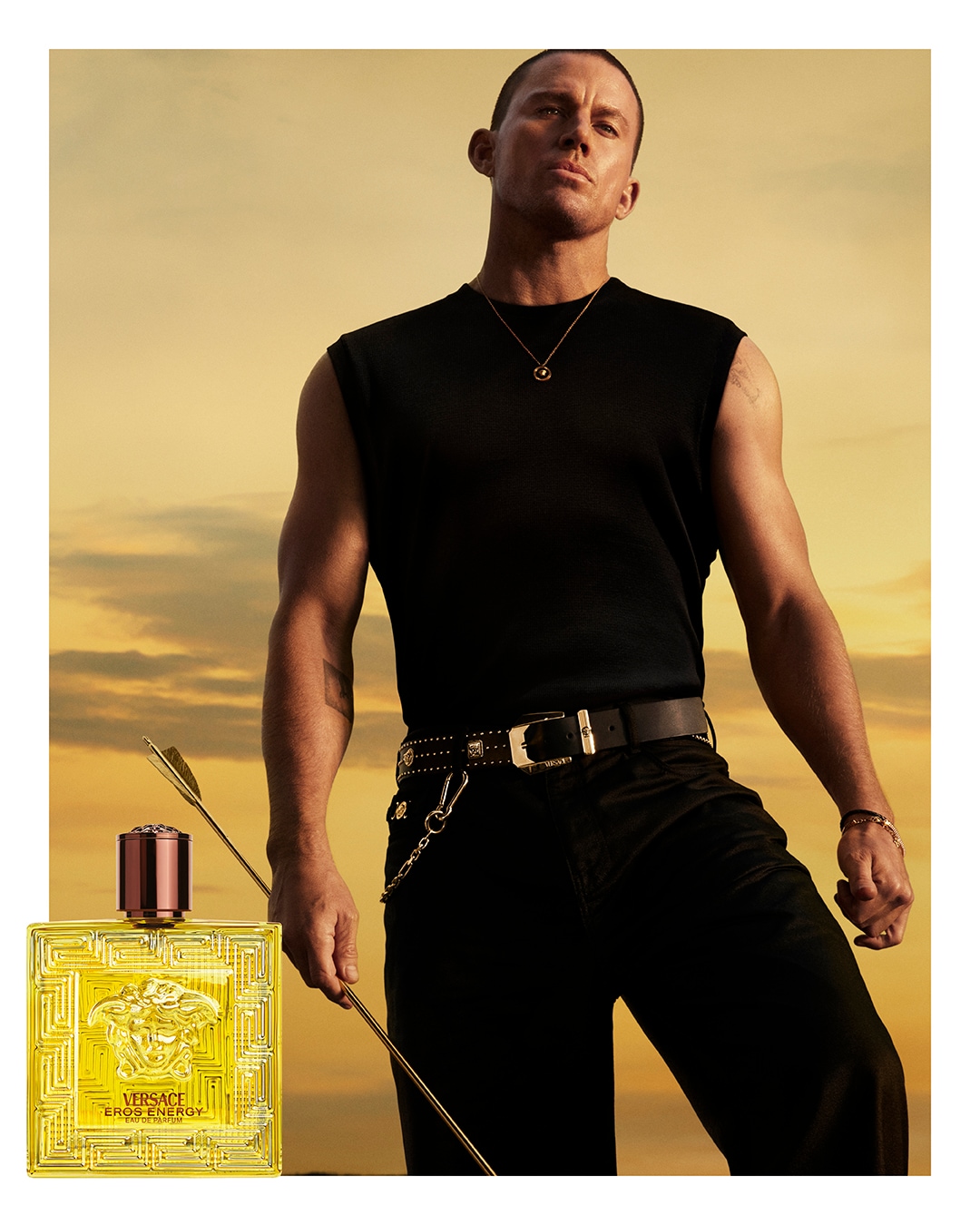 Mężczyzna w czarnym ubraniu pozuje z zapachem Versace Eros Energy, który ma żółty flakon ze złotą głową meduzy i brązową nakrętką.