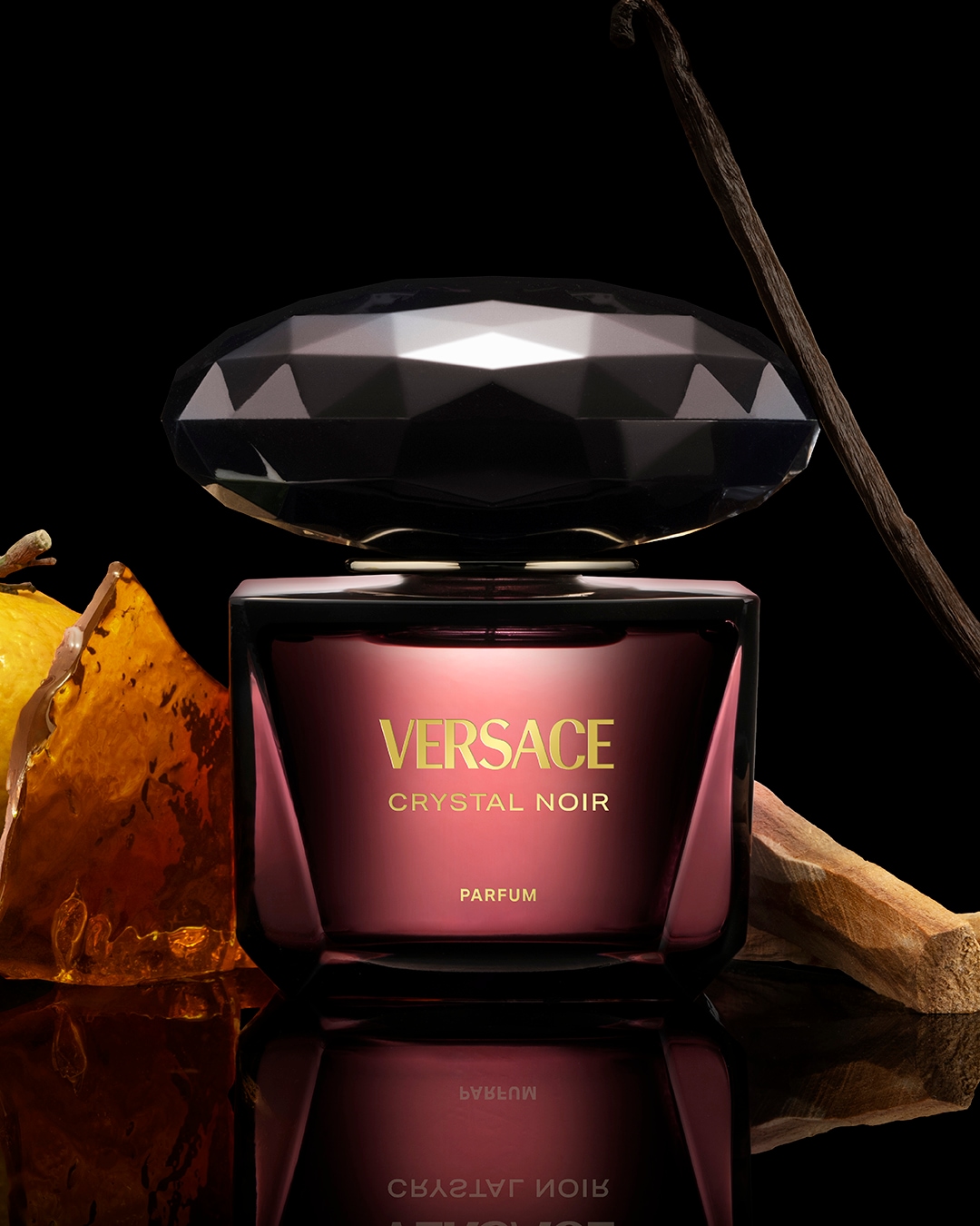 Butelka perfum Versace Crystal Noir o pojemności 90 ml ma ciemnoróżowy odcień i czarny kryształowy korek, a obok niej leżą laska wanilii i kawałek bursztynu.