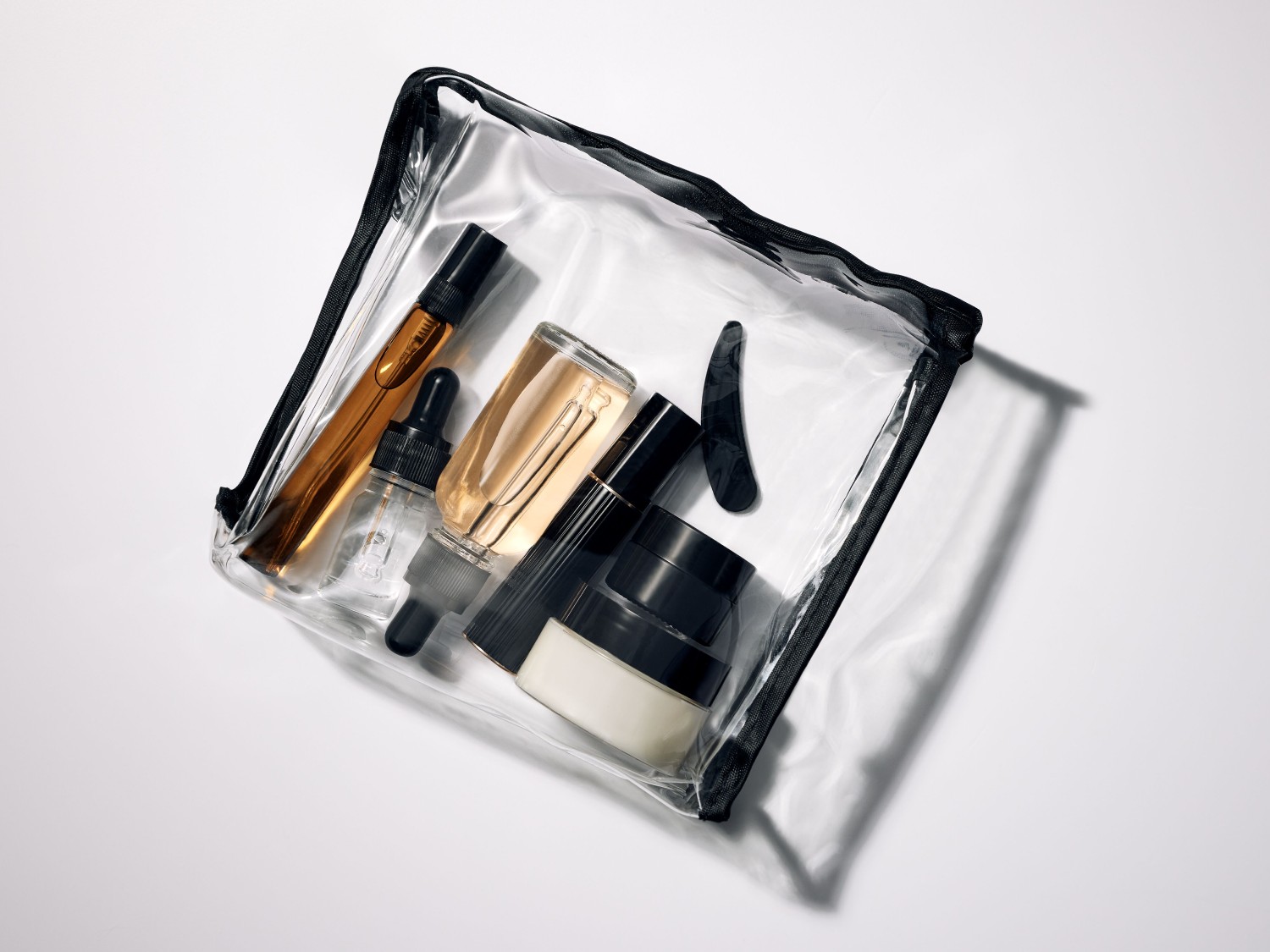 Een transparante make-up tas met zwarte rand bevat diverse huidverzorgingsproducten, waaronder flesjes met pipetten en crèmepotjes.
