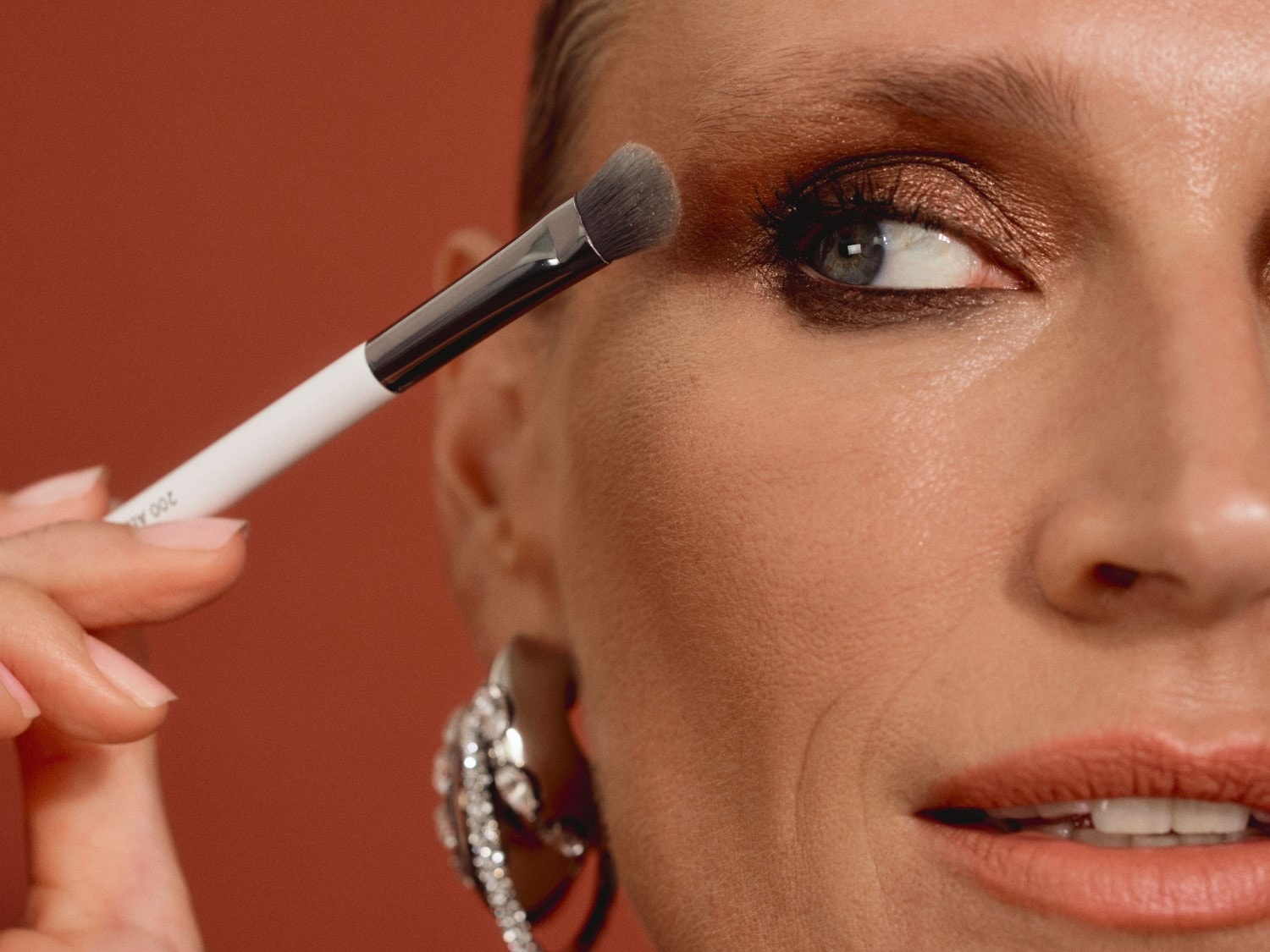 Een vrouw brengt oogschaduw aan met een make-up kwast, haar oog is opgemaakt in een glanzende bruine tint, en ze draagt opvallende zilveren oorbellen.