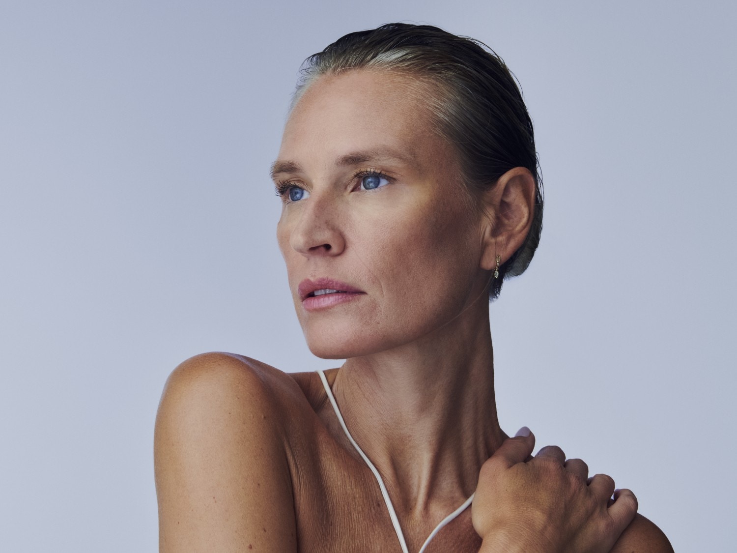 Porträt einer Frau mit hellblauen Augen, die ein weißes Oberteil und zarte Ohrringe trägt, während sie mit einer Hand ihre Schulter berührt und in die Ferne blickt.