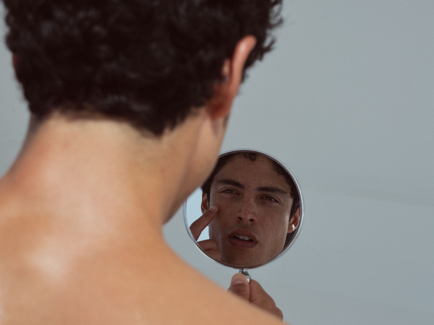 Ein Mann mit lockigem Haar betrachtet sein Gesicht in einem runden Spiegel und berührt mit einem Finger seine Wange.