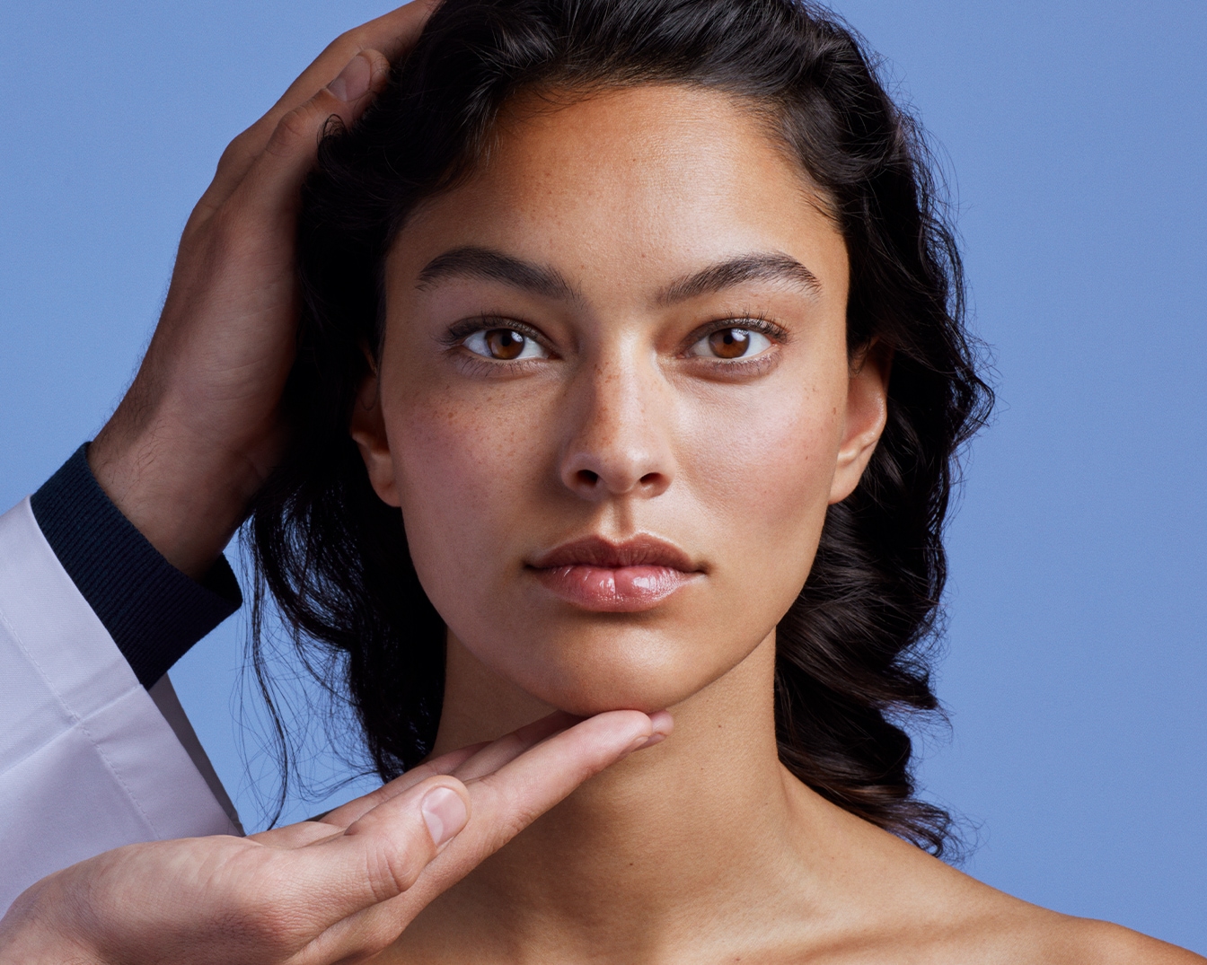 Primo piano di una donna con pelle abbronzata e occhi marroni, mentre un professionista esamina la sua pelle su sfondo blu.