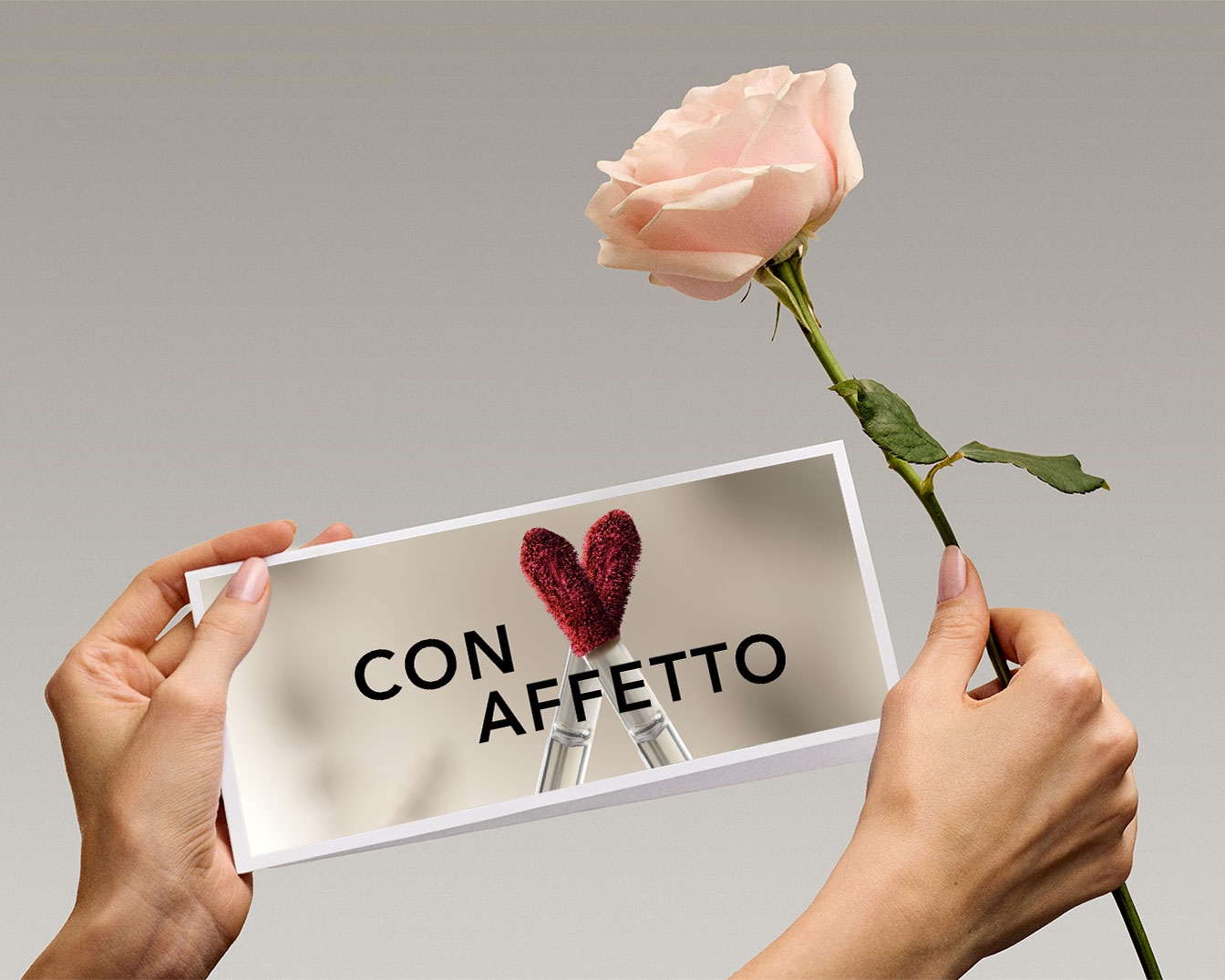 Una mano tiene una rosa color pesca accanto a una foto con la scritta 'CON AFFETTO' e due applicatori di lucidalabbra rossi a forma di cuore.