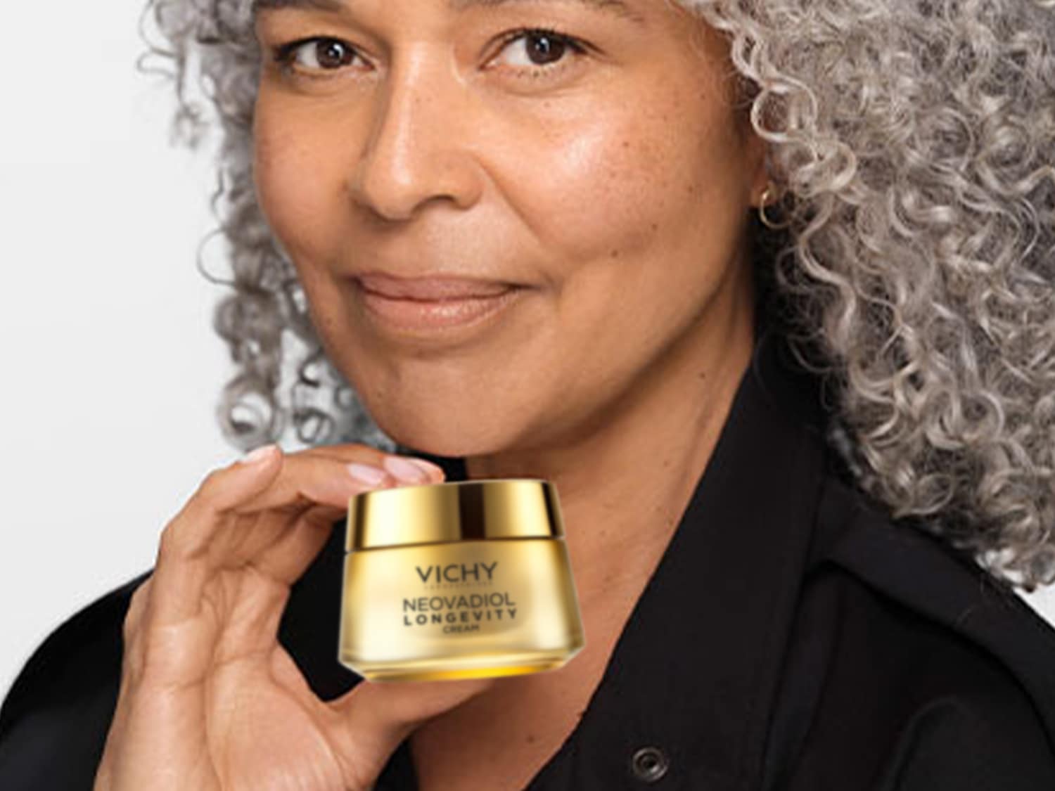 Una donna con i capelli grigi ricci tiene in mano un barattolo dorato di crema Vichy Neovadiol Longevity, promuovendo la cura della pelle e la bellezza.