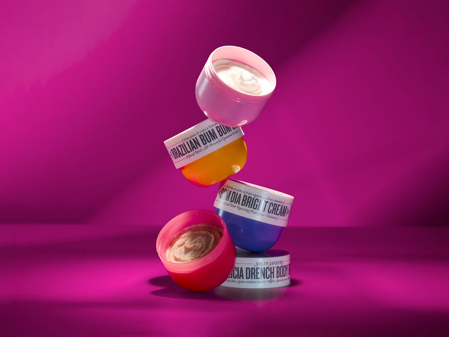 Découvrez l'ensemble de crèmes corporelles Sol de Janeiro, comprenant Brazilian Bum Bum, Bom Dia Bright Cream et l'hydratant Acai Drench Body, présentées dans des pots colorés empilés sur un fond rose vif.