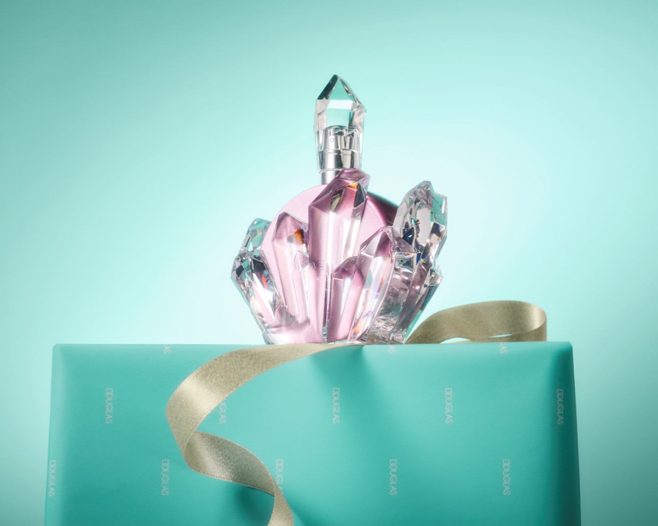 Il profumo rosa chiaro a forma di cristallo è adagiato su una confezione regalo turchese con un nastro dorato, il tutto su uno sfondo azzurro.