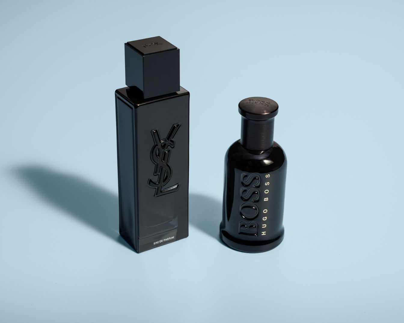 Twee zwarte parfumflessen, één van Yves Saint Laurent in een rechthoekige flacon en één van Hugo Boss in een ronde flacon, staan op een lichtblauwe achtergrond.